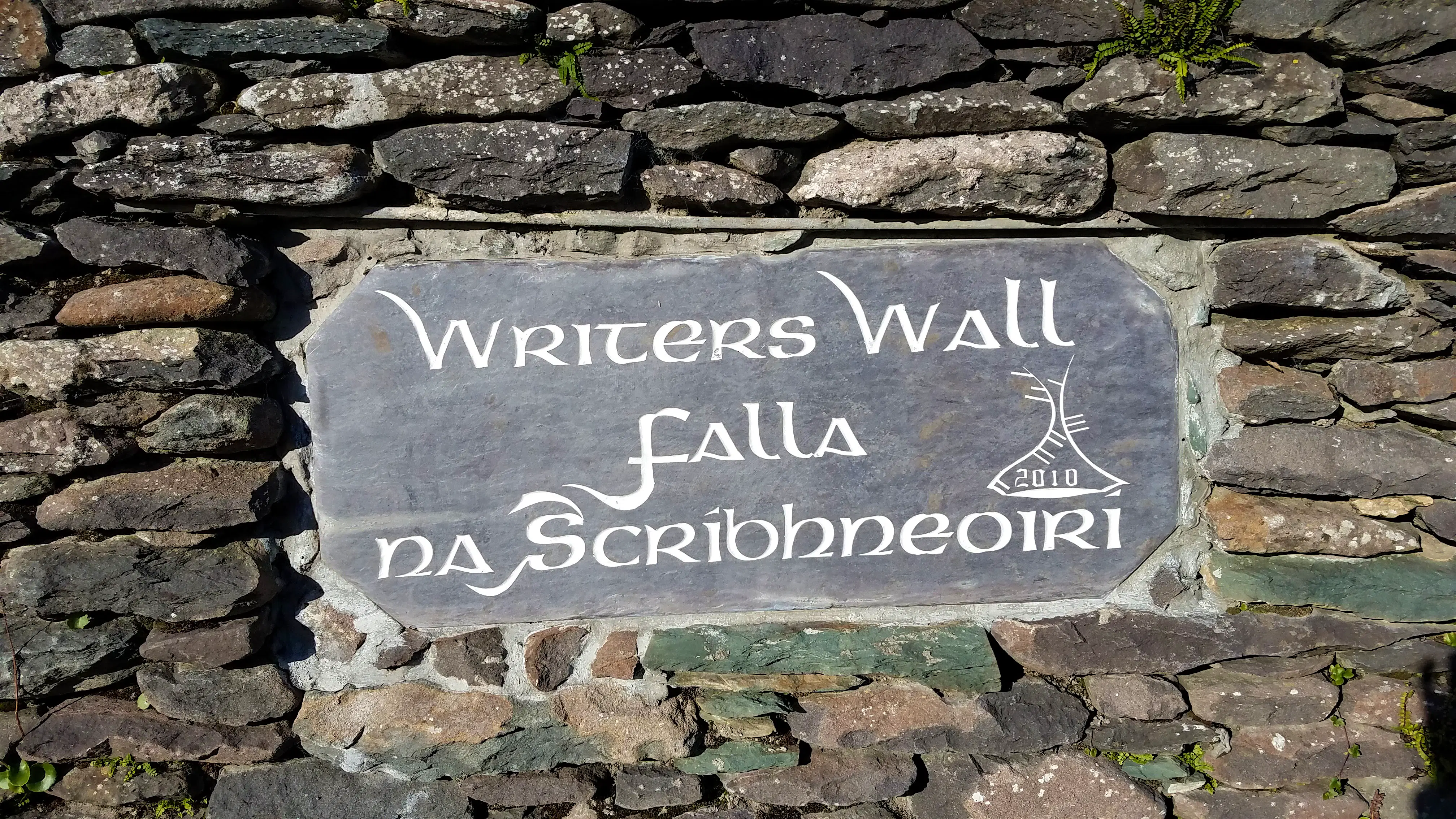 dingle-ireland-falla-na-scríbhneoirí.jpg A grey stone wall is set with a smooth, slate-colored stone plaque carved with a light grey text reading "Writers Wall Falla na Scríbhneoirí"
