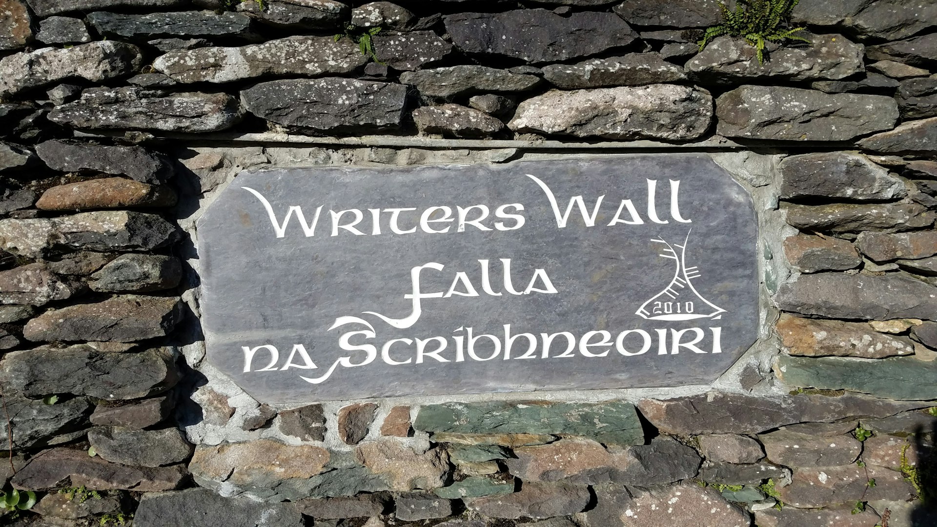 A grey stone wall is set with a smooth, slate-colored stone plaque carved with a light grey text reading "Writers Wall Falla na Scríbhneoirí"