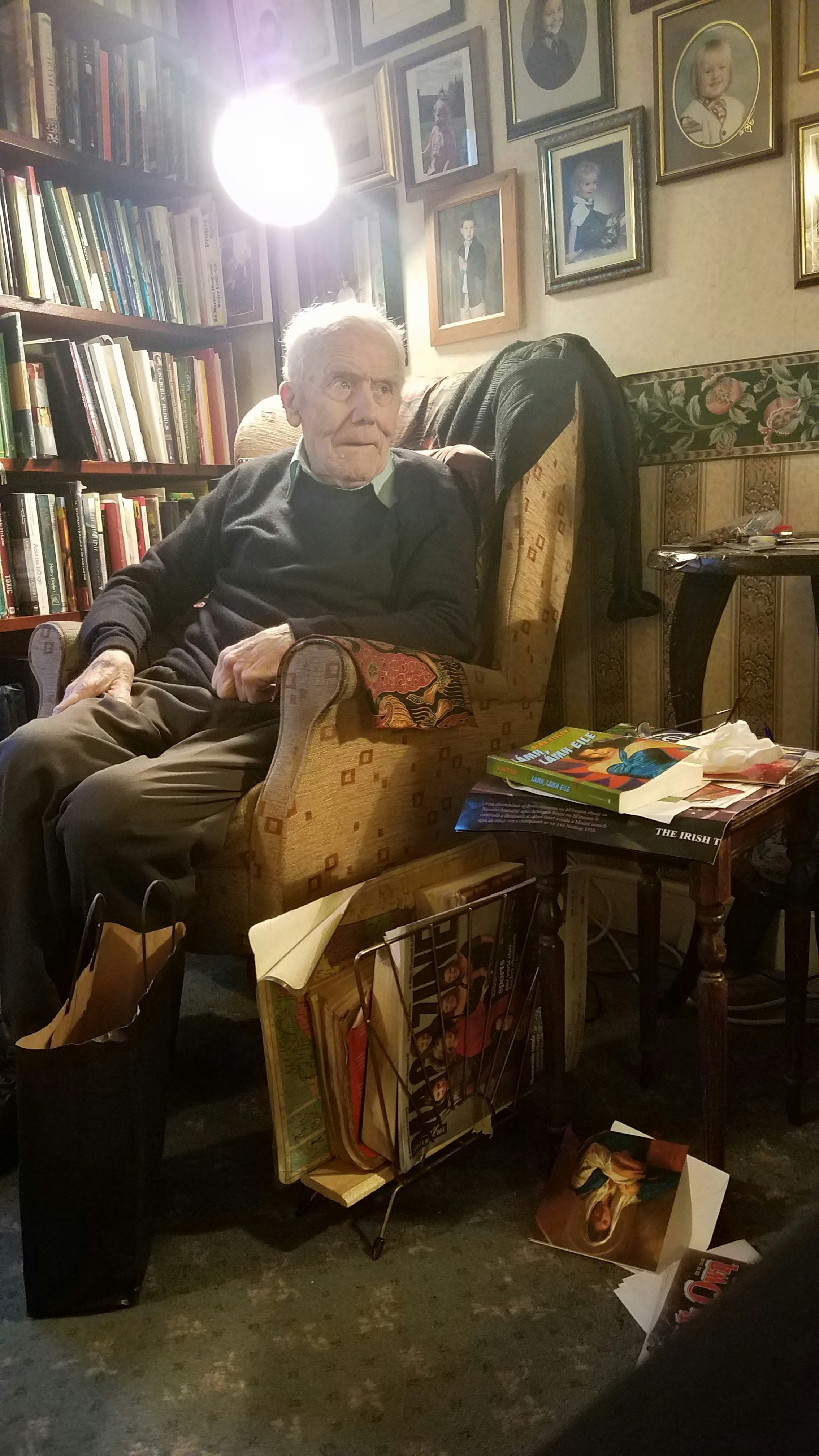 An elderly Irish man with white hair in a green collared shirt, dark green sweater, and olive slacks sits in a pale yellow wing back chair by a full bookcase under a wall of family portraits. The tables and magazine rack next to him and packed with books, periodicals, postcards, and other memorabilia. 