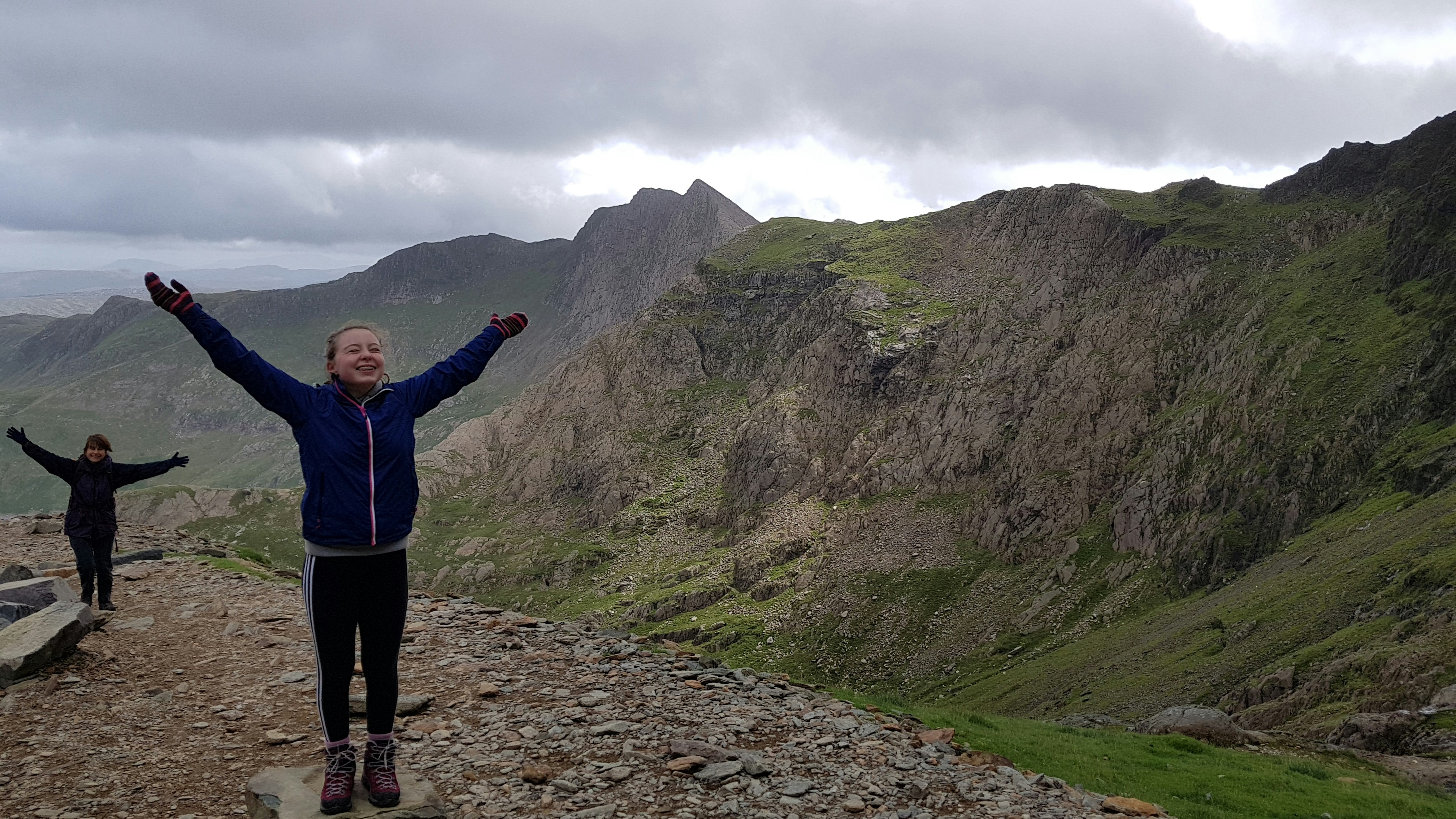 Tweens raise theri arms in enthusiasm at the top of Snowdon peak, the highest mountain in wales. Behind them, jagged sawtooth rock formations are coated in green moss against a grey cloudy sky..jpg