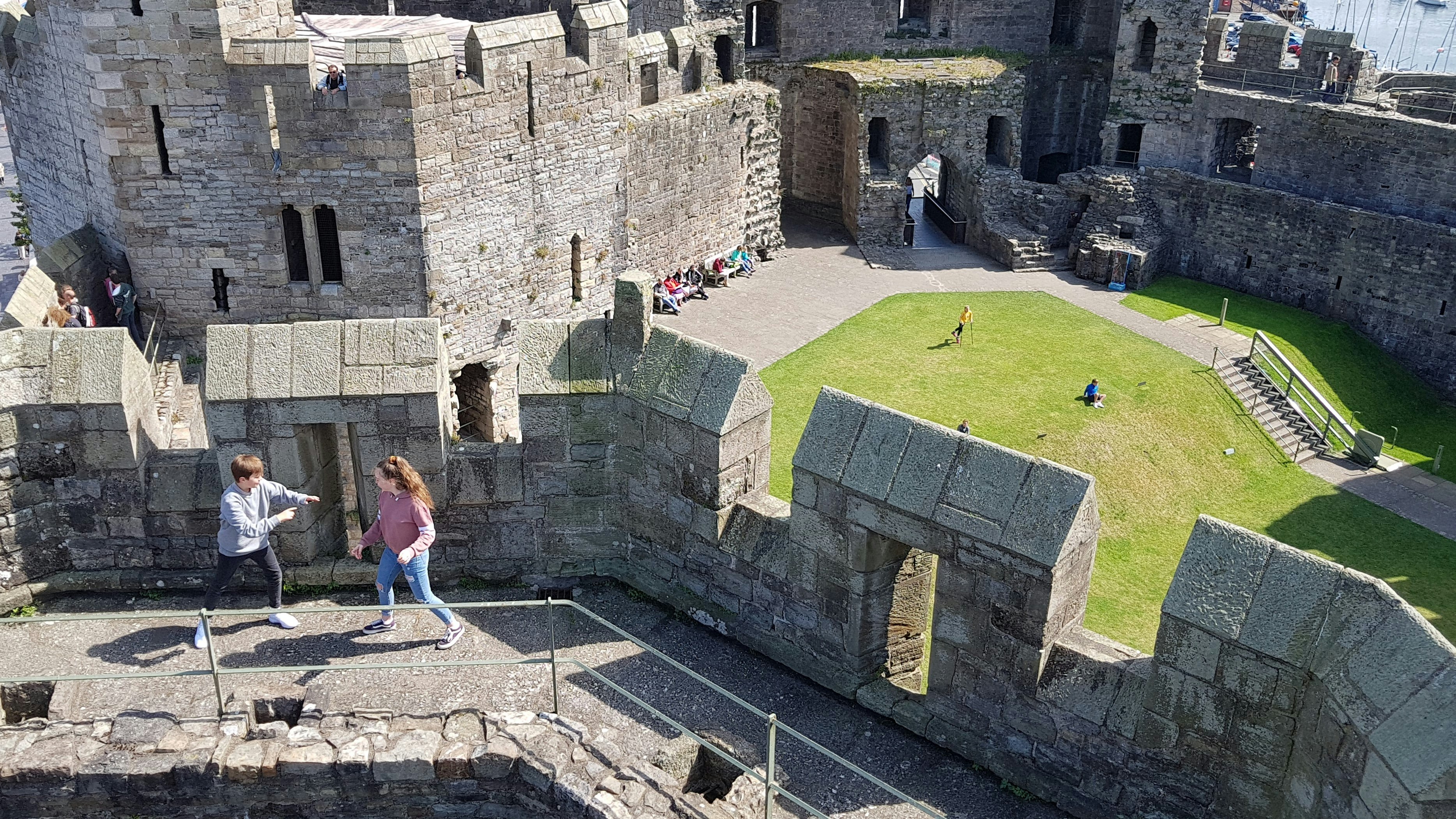 Two tweens in jeans and sweatshirts pretend to spar near stone crenelations at Caernarfon Castle in Wales. Behind them is a green lawn far below where more families play and explore. In the very far background to the right, you can just make out the masts of ships behind the castle. 