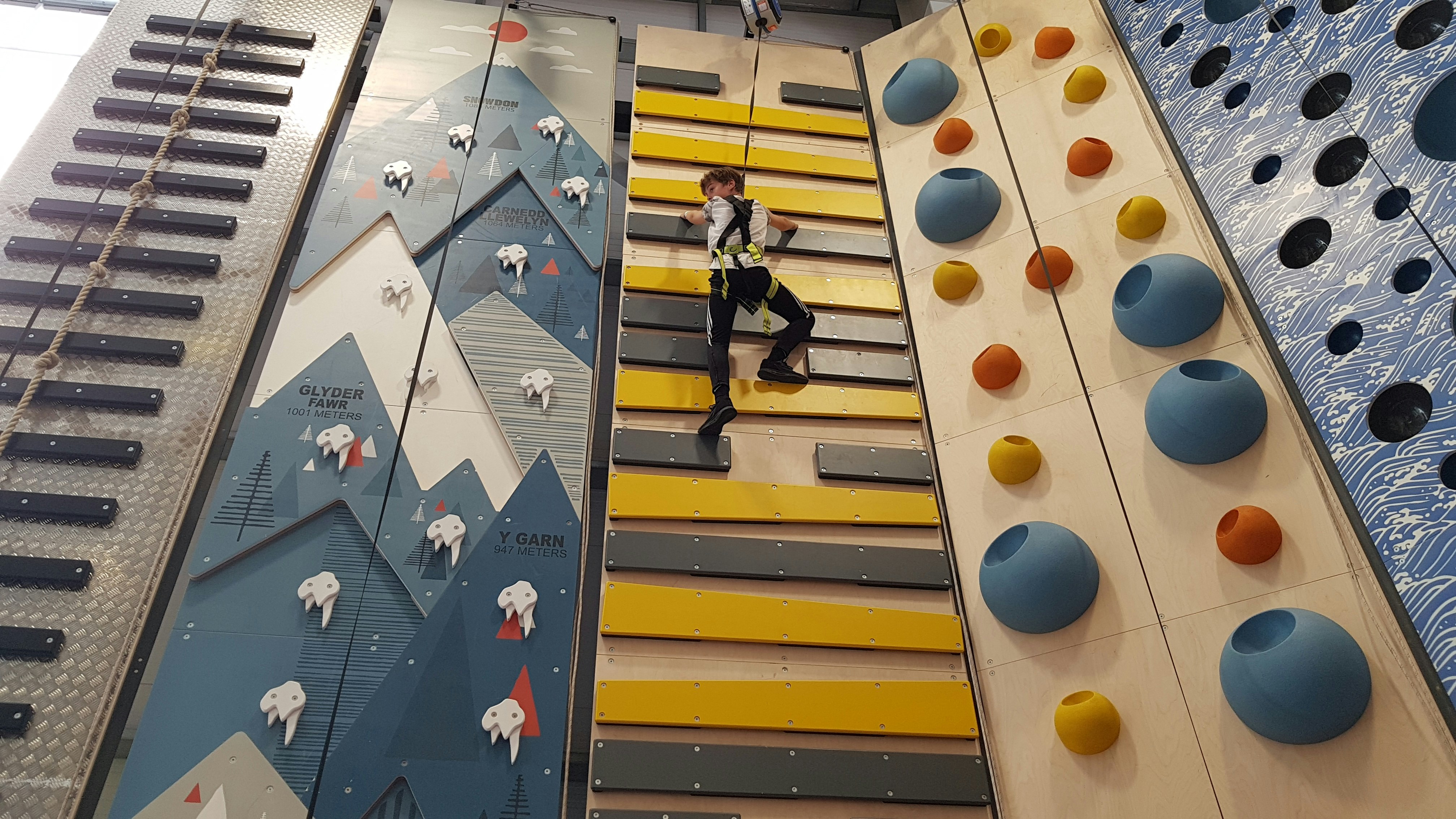 A pre-teen boy in black track pants and a white shirt with a climbing harness and belay rope on climbs yellow and black planks next to a more technical blue and white climbing wall designed to look like jagged mountain peaks.