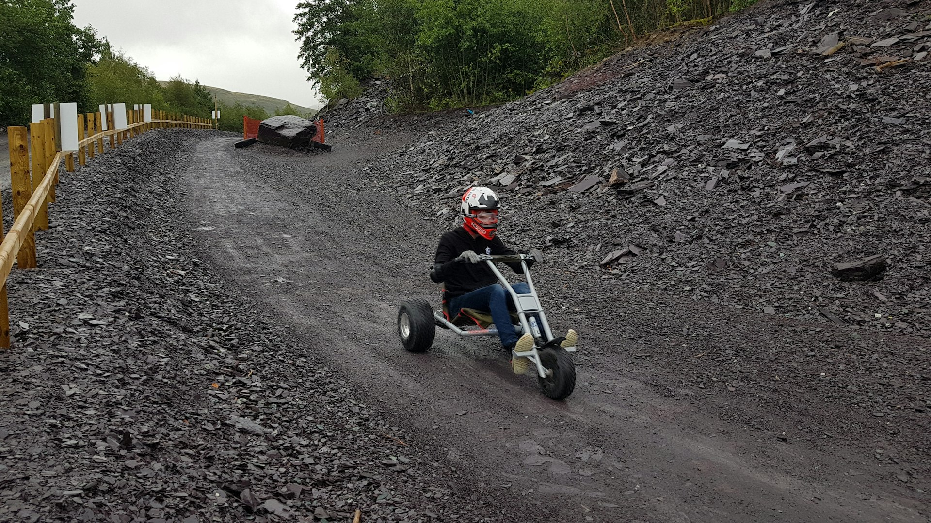 A pre-teen boy in a black sweater and white and red motorcycle helmet rides down a black shale track on a three-wheeled steel racing trike
