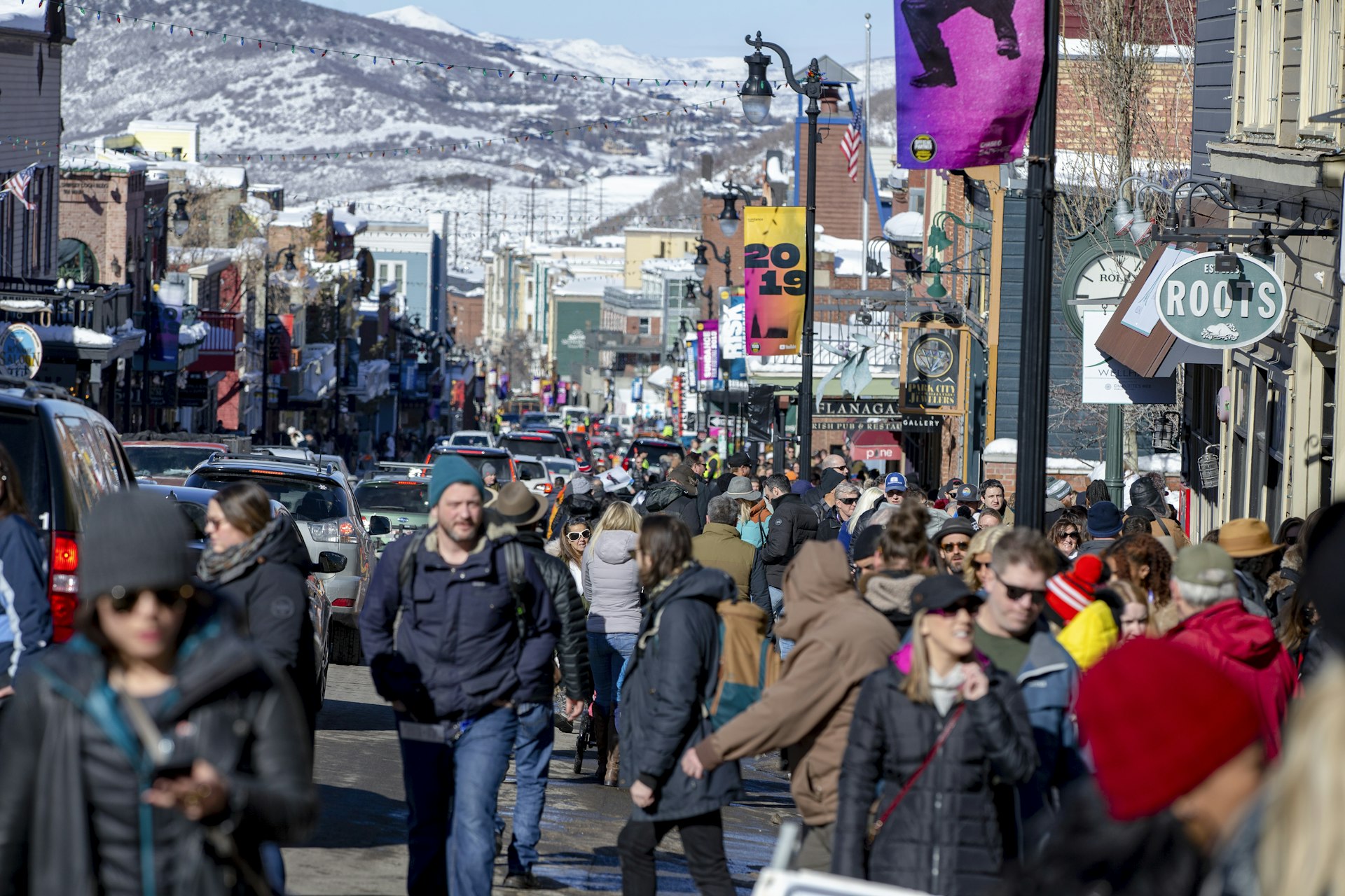 A crowd of Sundance festival goers in winter goats, hats, and sunglasses crowd Main Street in Park City, where the lampposts have yellow and pink banners reading 2019 for last years festival