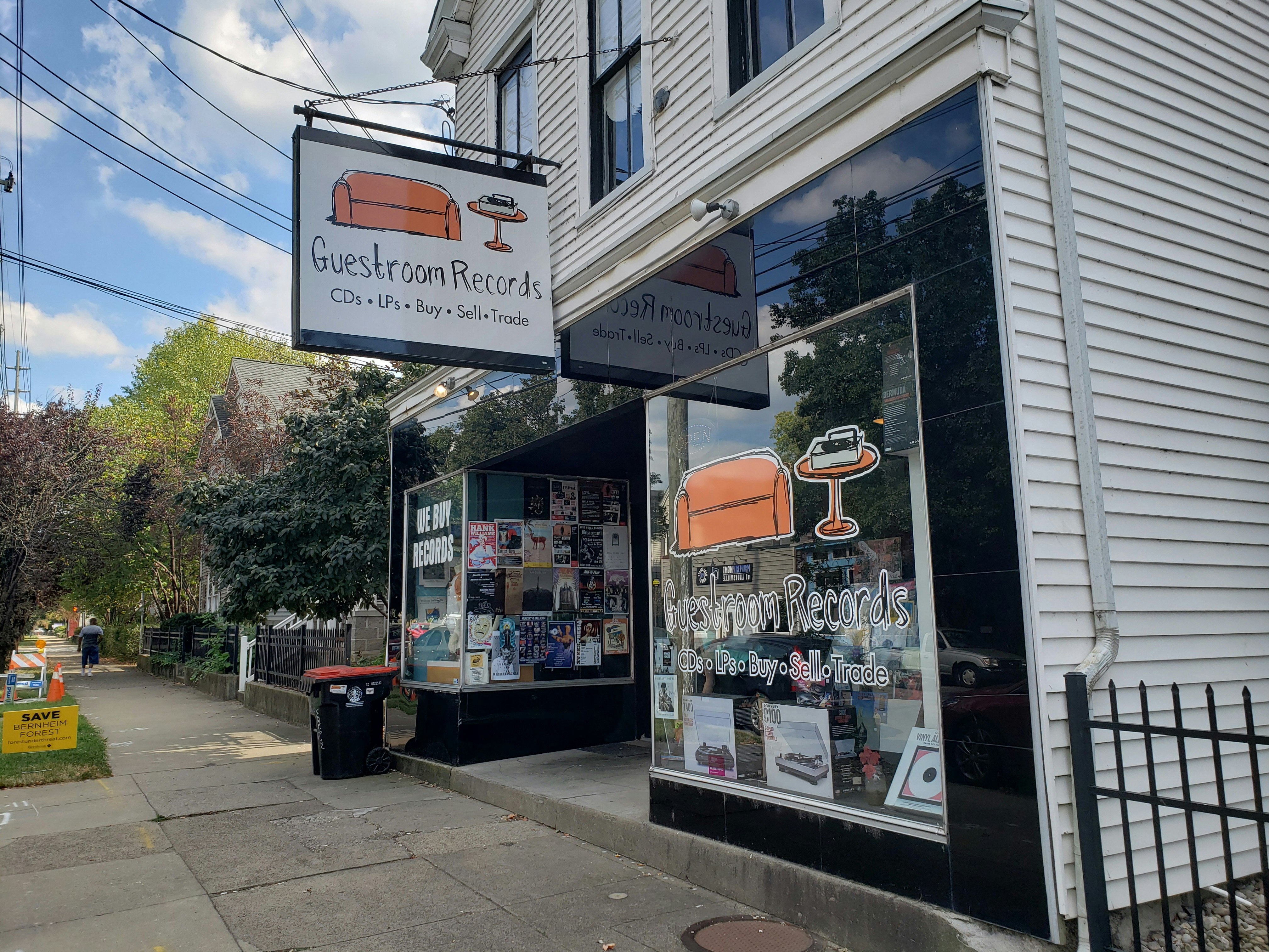 The exterior of Guestroom Records in Louisville, Kentucky is a two-story building with white clapboard siding. The storefront consists of two plate-glass windows on either side of a doorway, filled with colorful event posters and a large cartoony logo of an orange couch, an orange side table, with a record player on top. The concrete sidewalk extends to the left, showing a pretty tree-lined street and a pedestrian. 