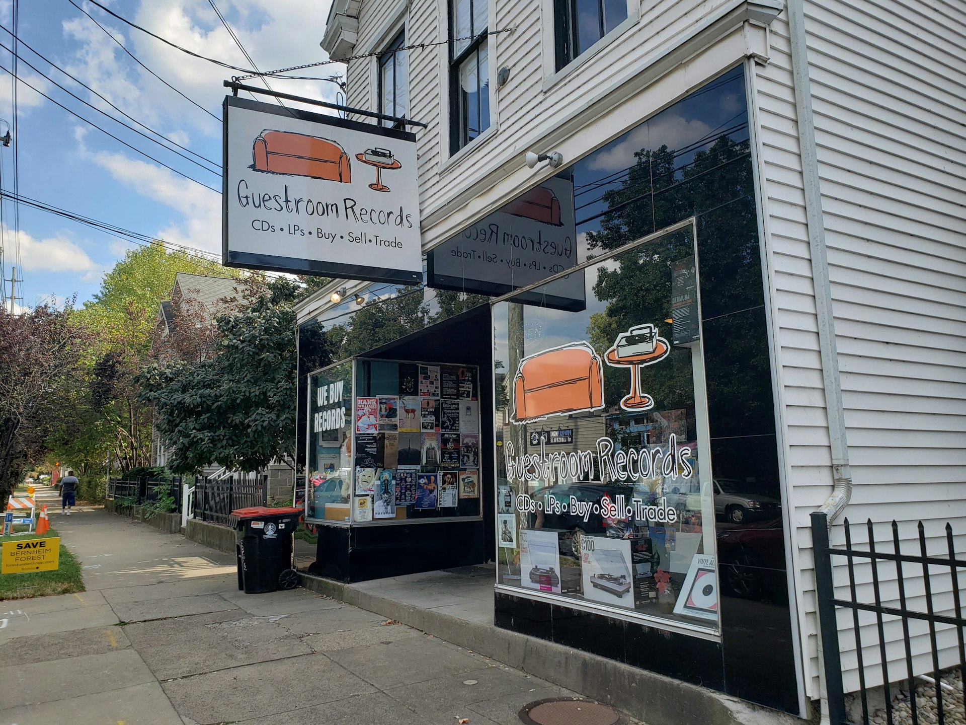 The exterior of Guestroom Records in Louisville, Kentucky is a two-story building with white clapboard siding. The storefront consists of two plate-glass windows on either side of a doorway, filled with colorful event posters and a large cartoony logo of an orange couch, an orange side table, with a record player on top. The concrete sidewalk extends to the left, showing a pretty tree-lined street and a pedestrian. 