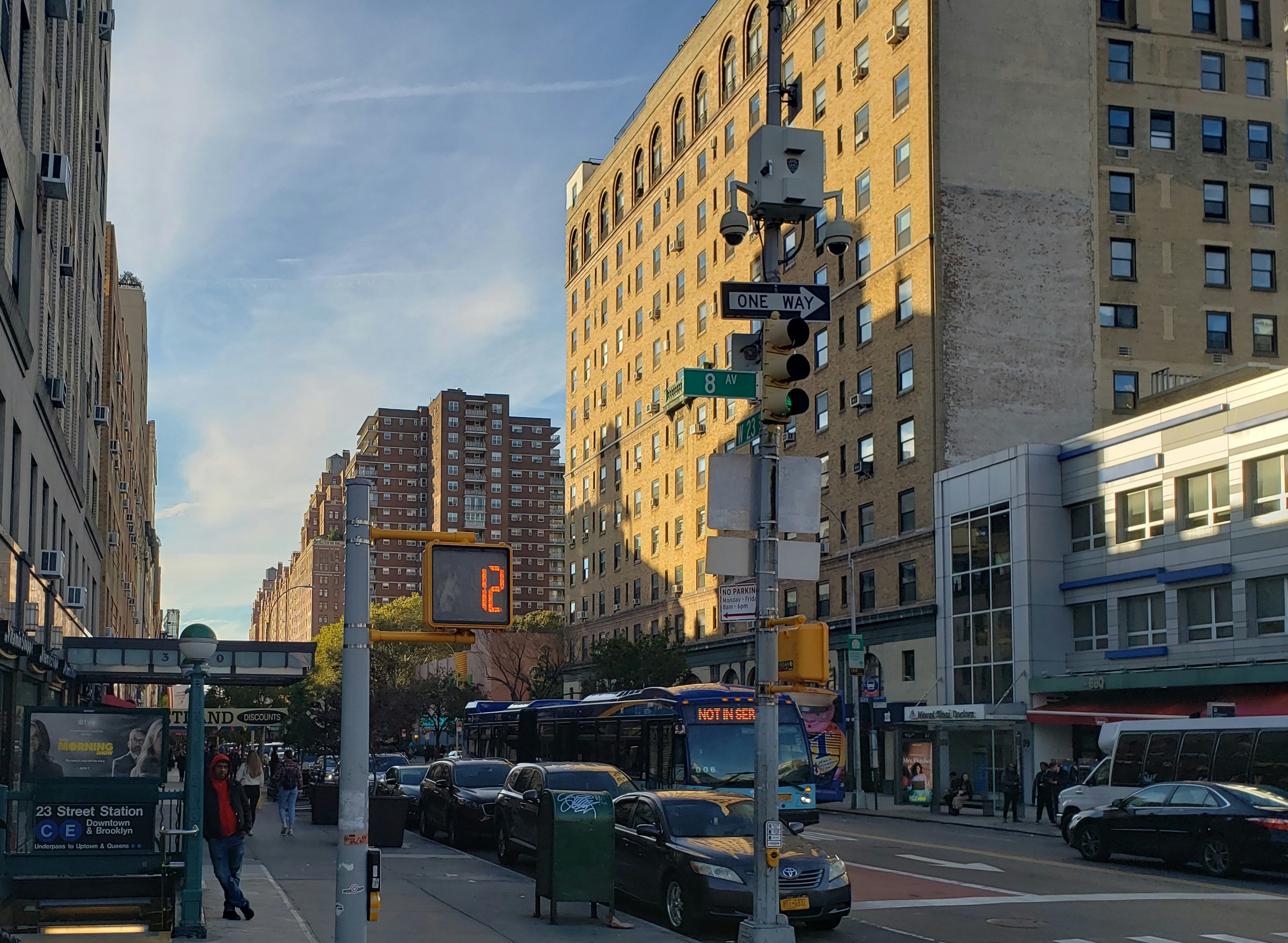 The yellow brick buildings at the corner of 23rd and 8th in New York City glow golden in the afternoon light, overlooking a subway station where a man is leaning on the railing, an out-of-service blue bus, and a row of black cars parallel parked on the street