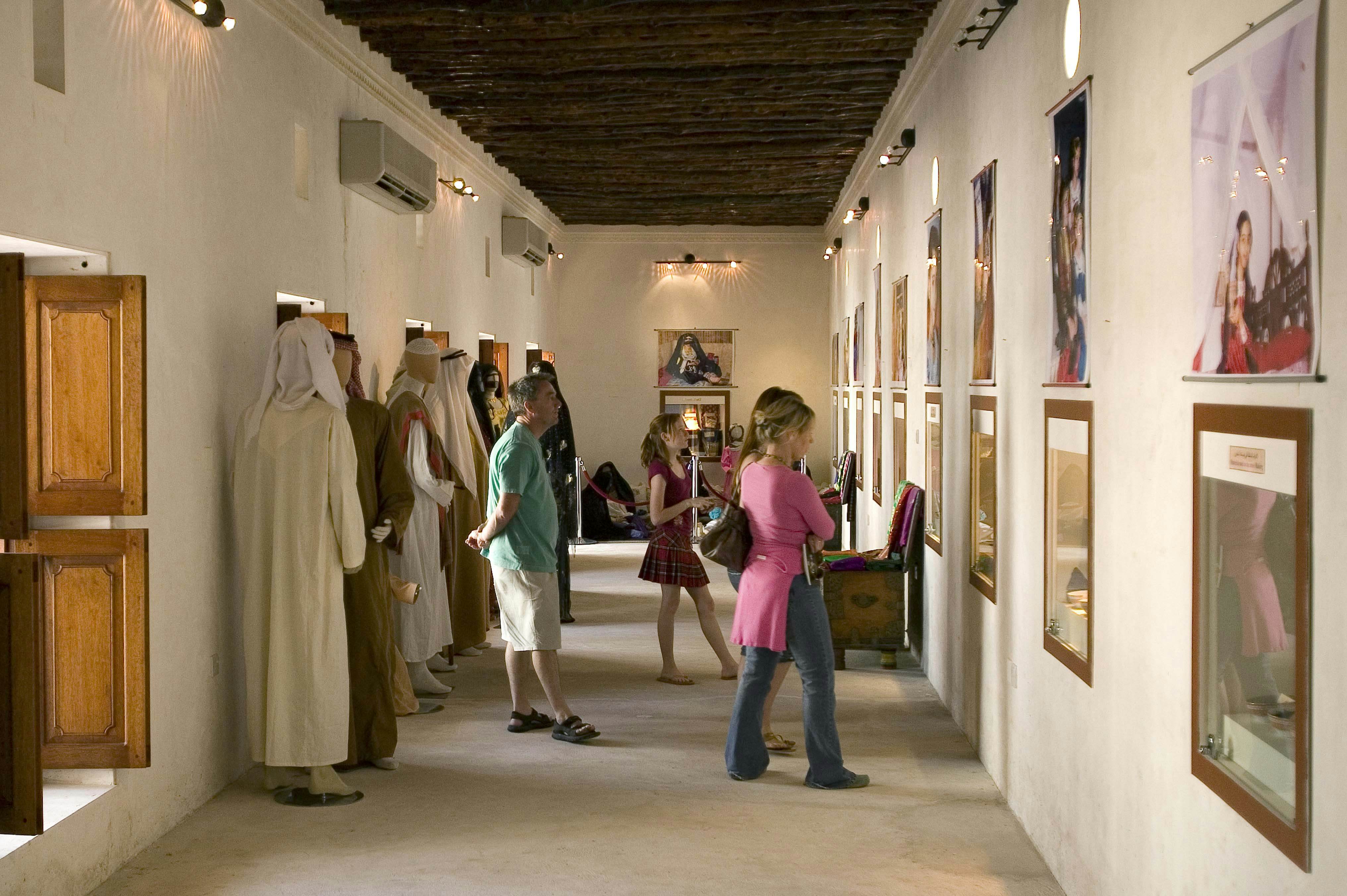 A family are looking at displays on the wall in a long, white-walled room in a museum. Behind them, several mannequins are adorned in traditional Emirati dress.