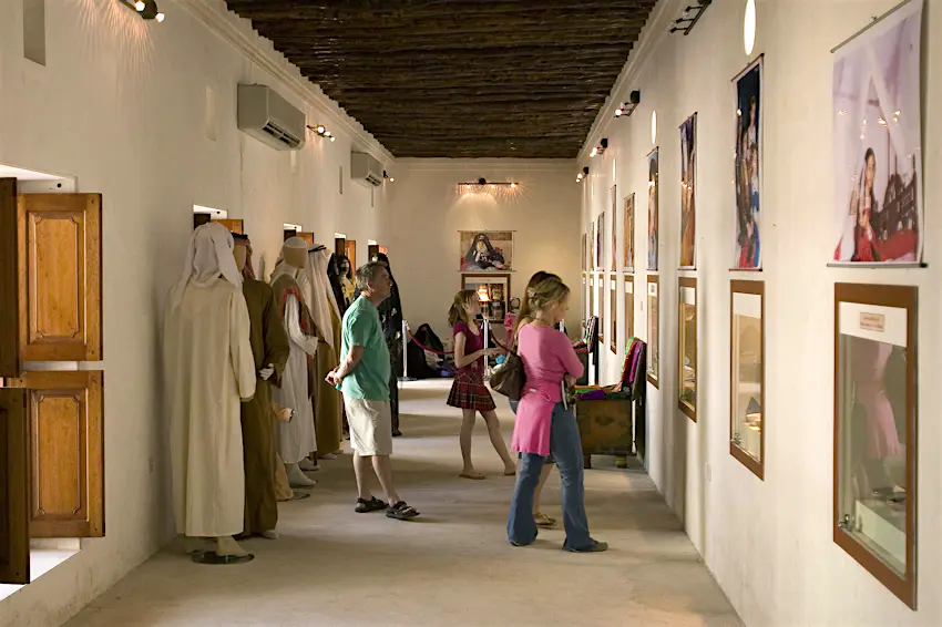 heritage_museum_sharjah.jpg A family are looking at displays on the wall in a long, white-walled room in a museum. Behind them, several mannequins are adorned in traditional Emirati dress.