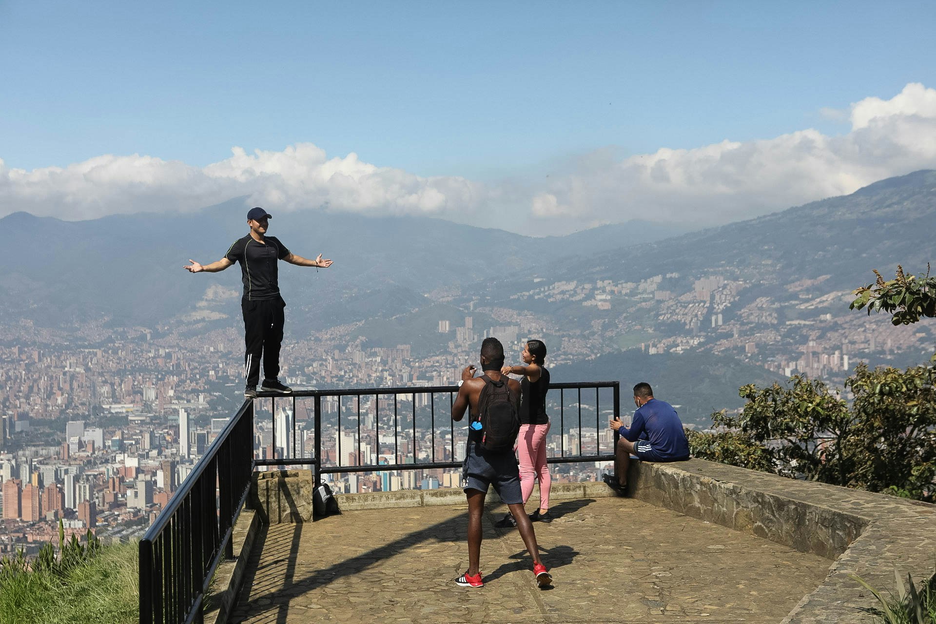 A man in black track pants and a athleisure shirt and black ball cap poses by standing on the corner of a railing at an overlook that offers panoramic views of Medellin below.jpg