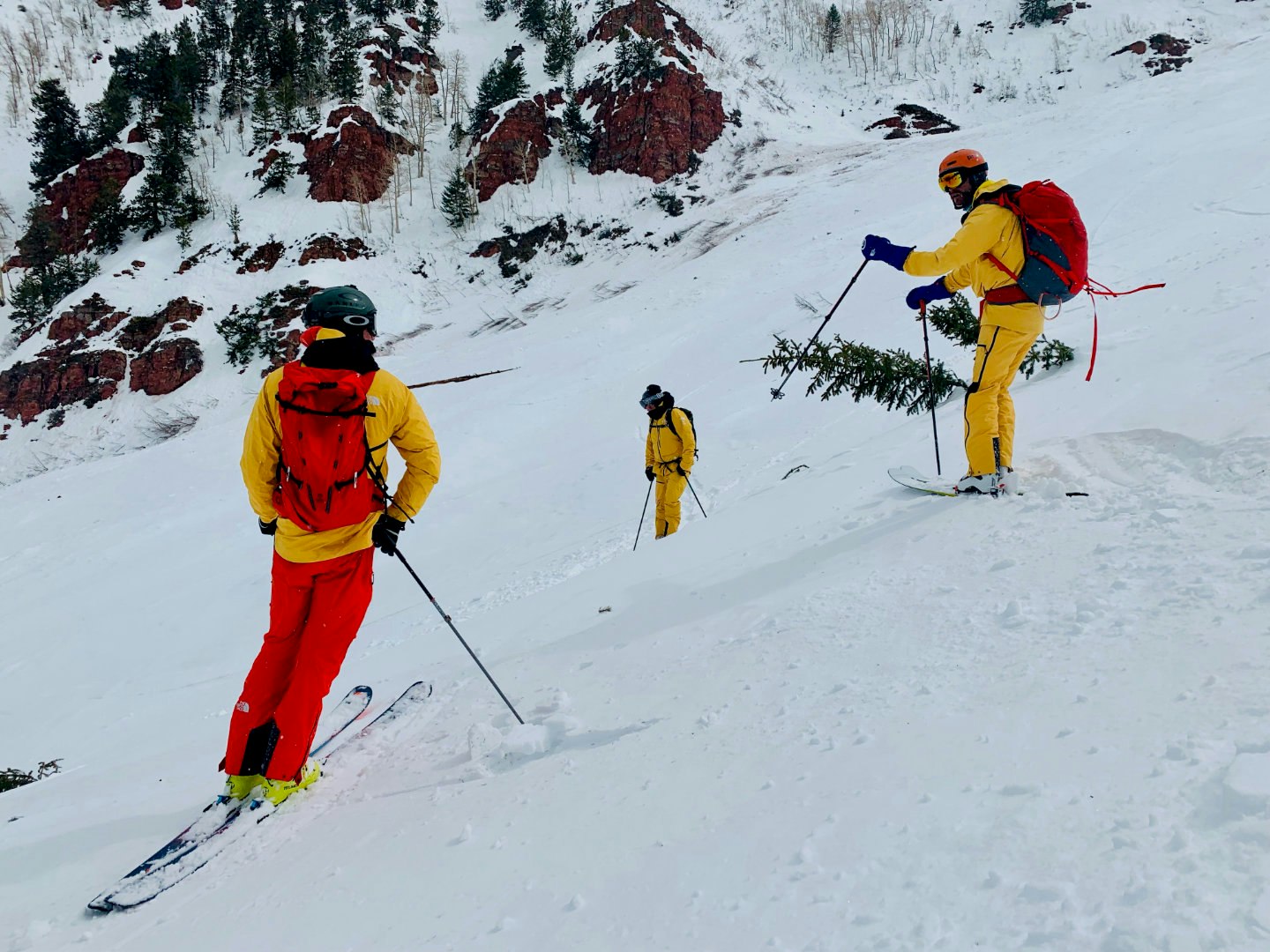 Three skiers descend a slope in Aspen thickly covered with snow. All are wearing yellow and red FutureLight pants and jackets from North Face. In the background are large boulders and trees. 