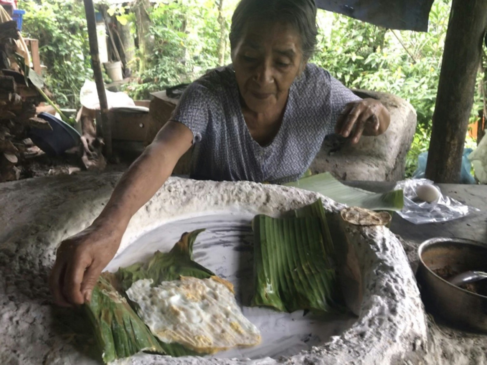 A woman leans over a stone structure as she prepares to fold a banana leaf over fried eggs; Xantolo