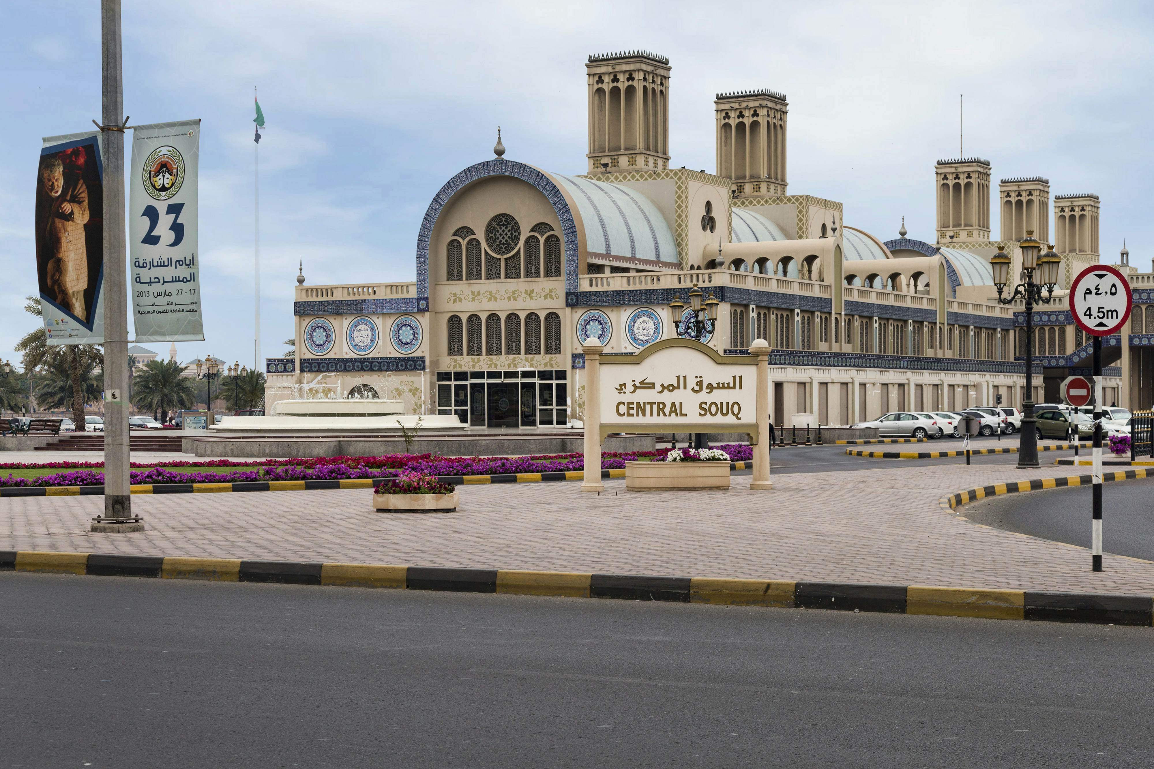 A wide-angle shot of a huge sandy-coloured building which is accented with decorative blue tiles. Outside is a sign that reads "Central Souk" in both Arabic and English.