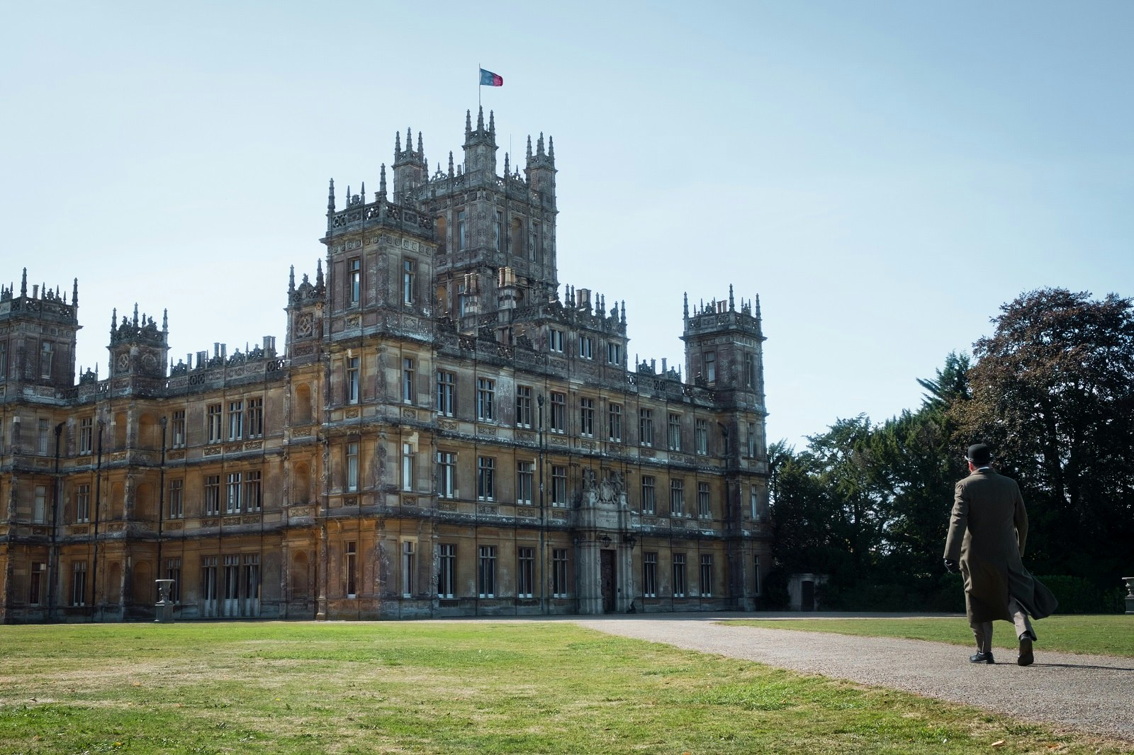 A man in a long beige coat and a bowler hat strides down the path towards Highclere Castle, which is the filming location for Downton Abbey.
