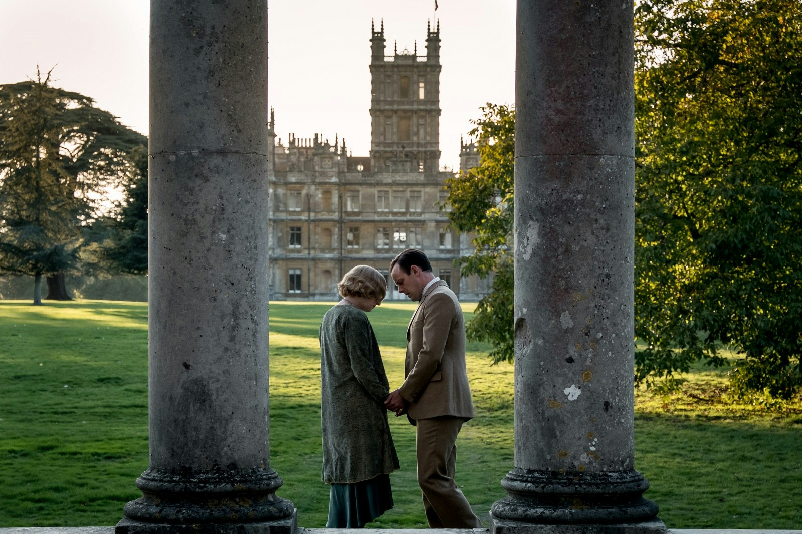 A man and a woman stand holding hands with their heads hanging low. Highclere Castle, doubling as Downton Abbey, can be seen in the background.
