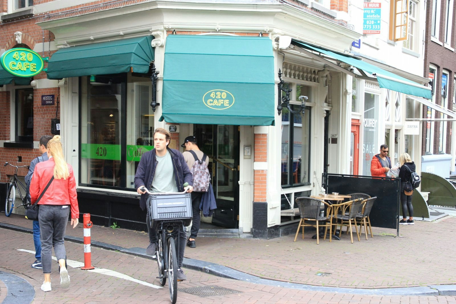 The exterior of 420 Coffeeshop, Amsterdam. People are cycling and walking by. The door of the shop has a green covering featuring the logo and there is covered outdoor seating. 