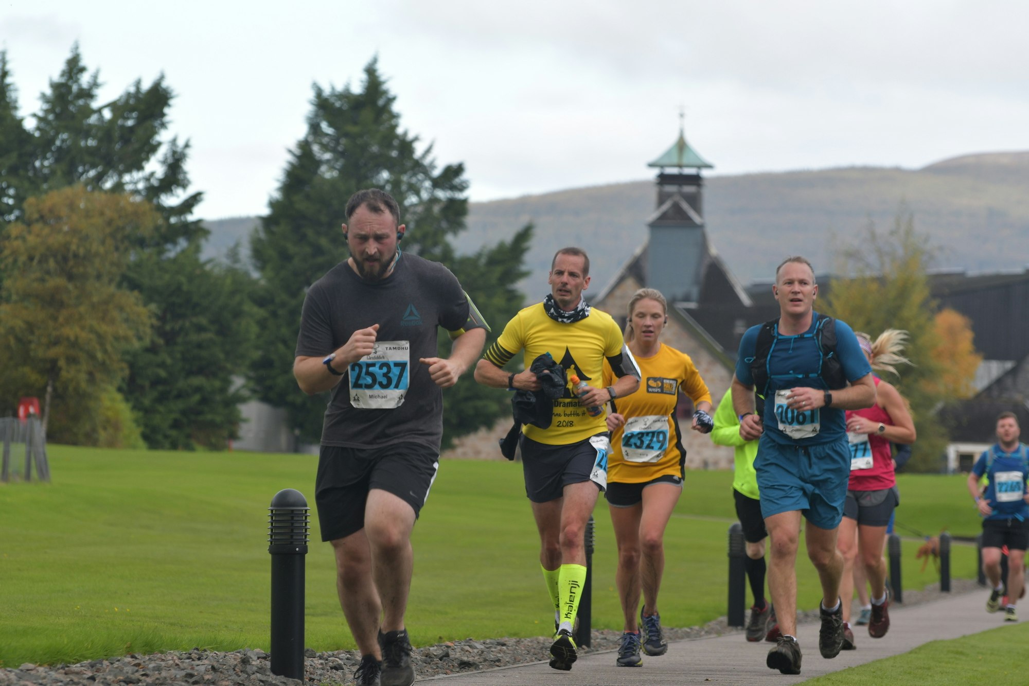 A group of racers are running along a path through landscaped grounds on an overcast day. Behind them, the turret of a distillery is visible.