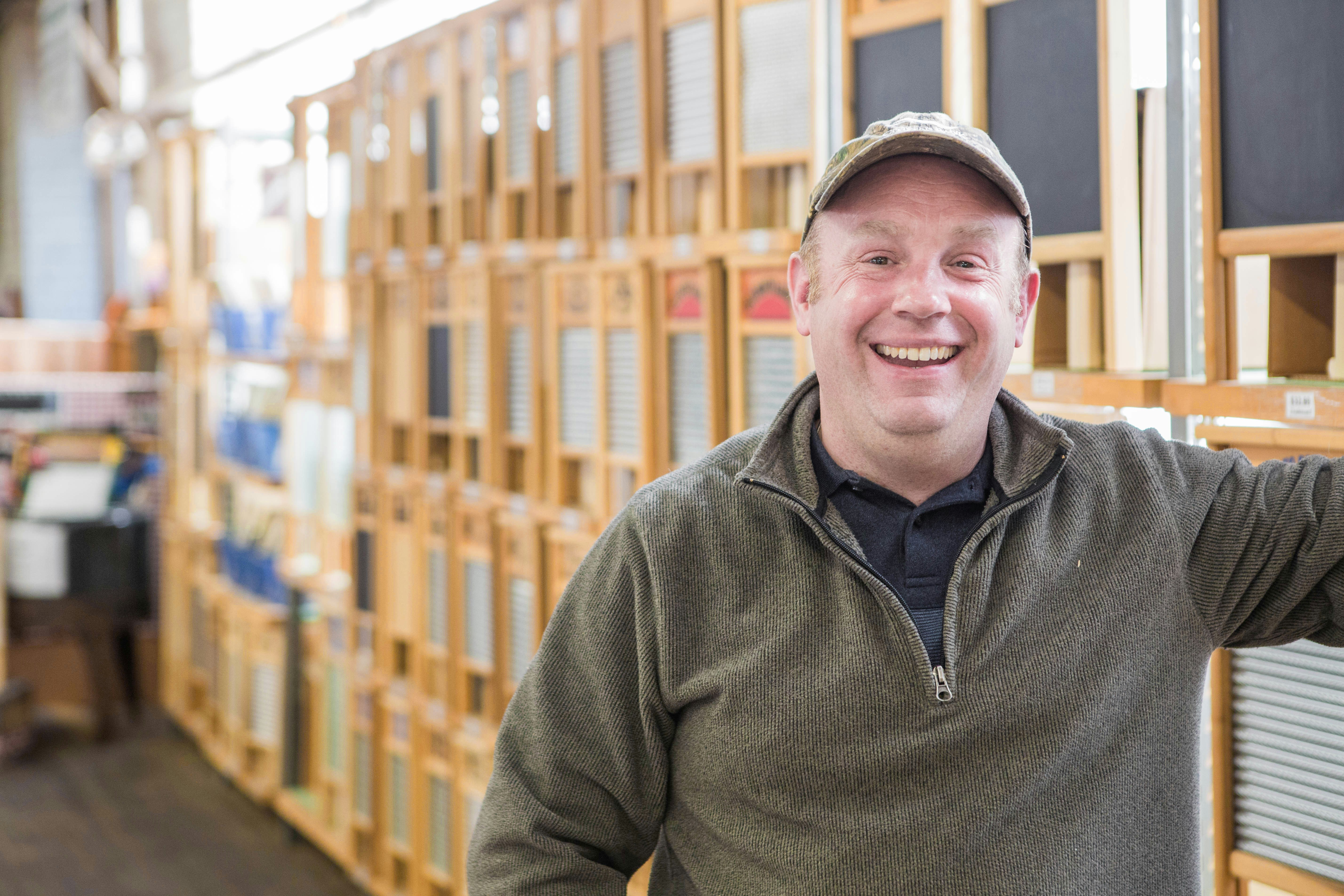 James Martin, co-owner of the Columbus Washboard Company, stands with a big cheerful smile in front of a row of washboards. He wears an olive green quarter-zip pullover with a grey polo shirt underneath and a tan ball cap. 
