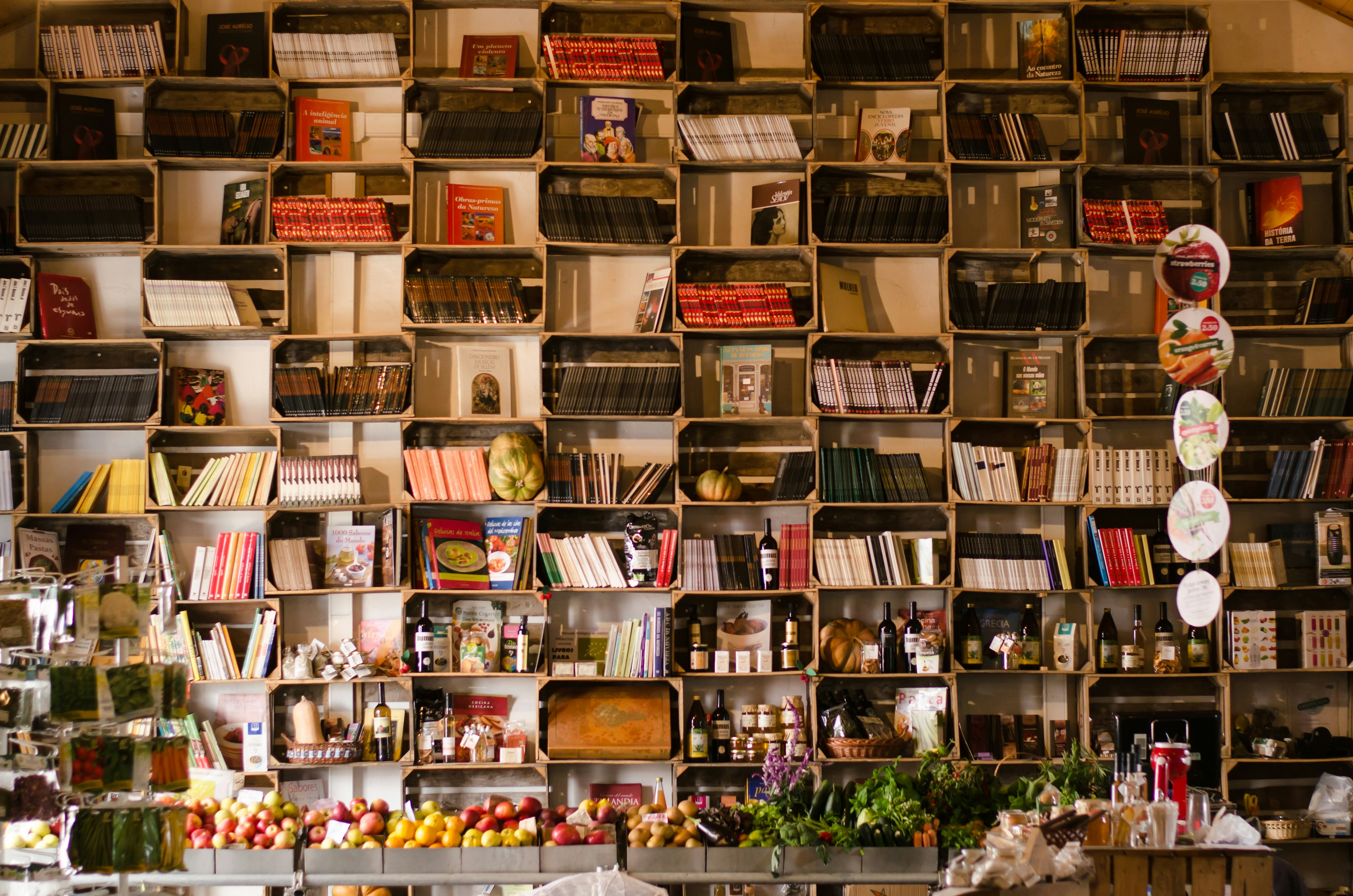 A wall of compartmentalised bookshelves is filled with books; some shelves also have wine bottles and fresh squashes. A table of inviting fruit and vegetables sits underneath the books.