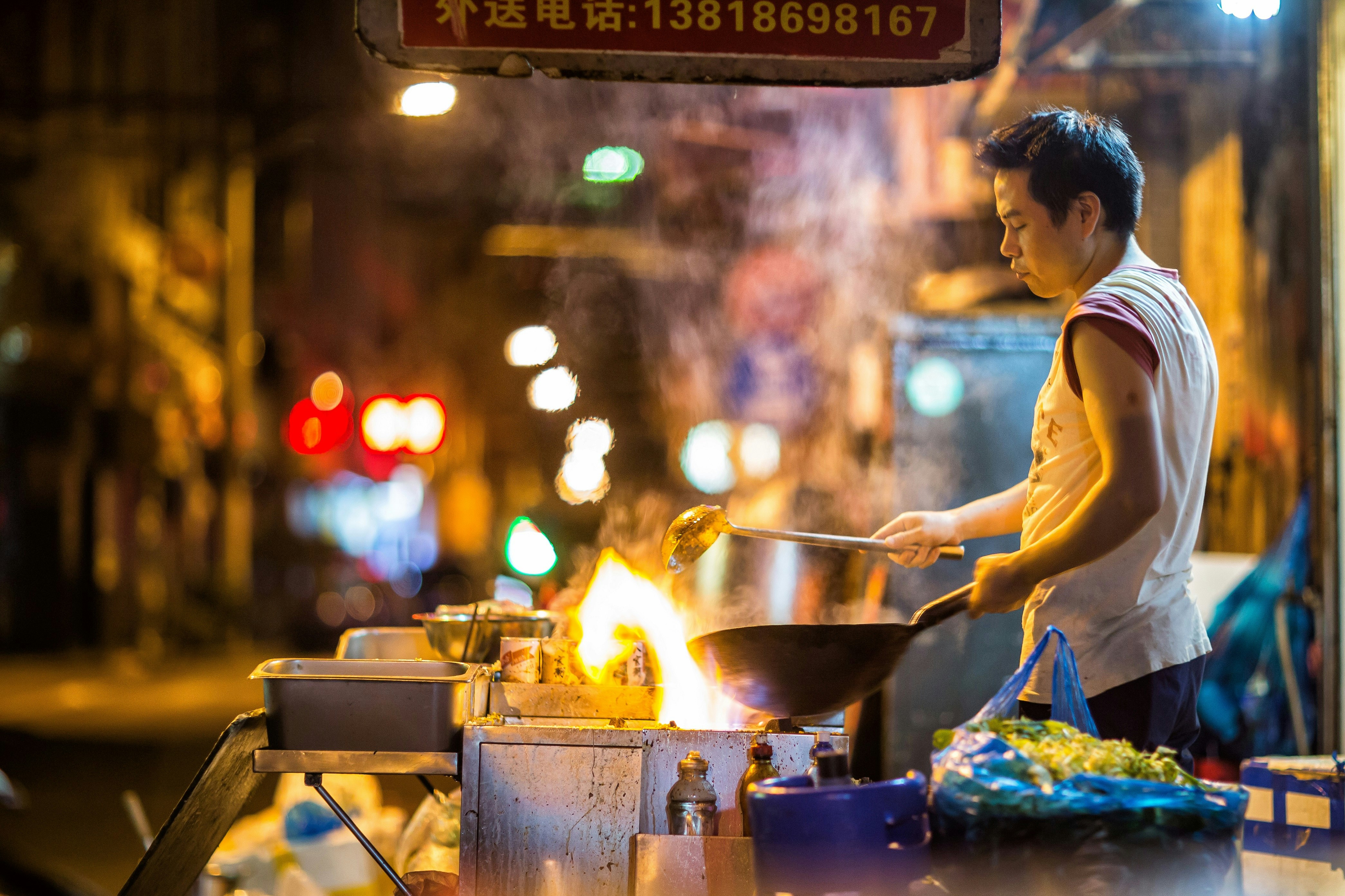 A vendor cooks food in a wok over an open flame on the darkened evening streets of Shanghai. The vendor is in sharp focus while the background is blurred.