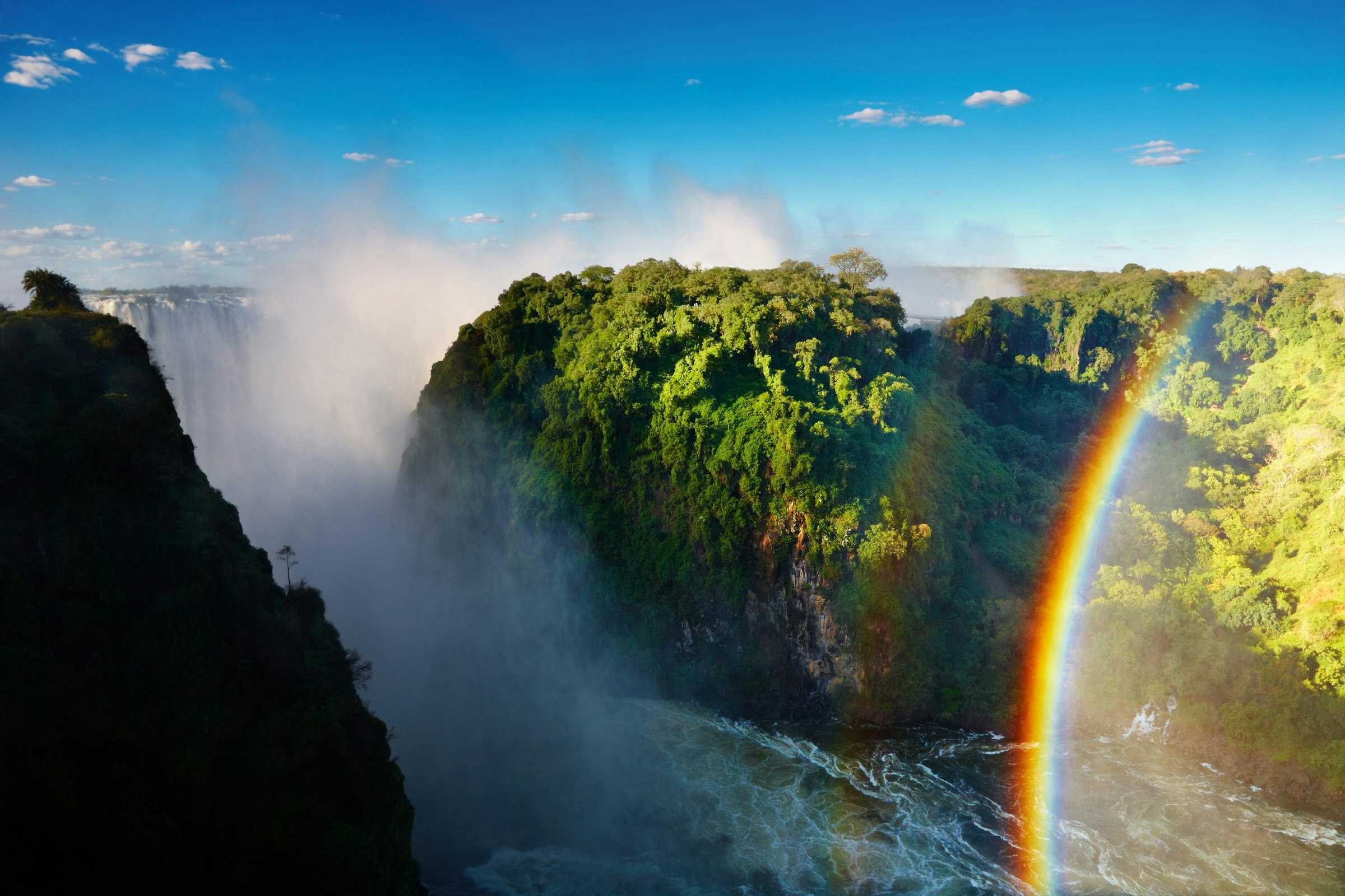Between a silhouetted rocky cliff and rainforest-clad outcrop is a seething Zambezi; a arching rainbow bills the right of the image, while the Victoria Falls are seen 'smoking' between the two cliffs.