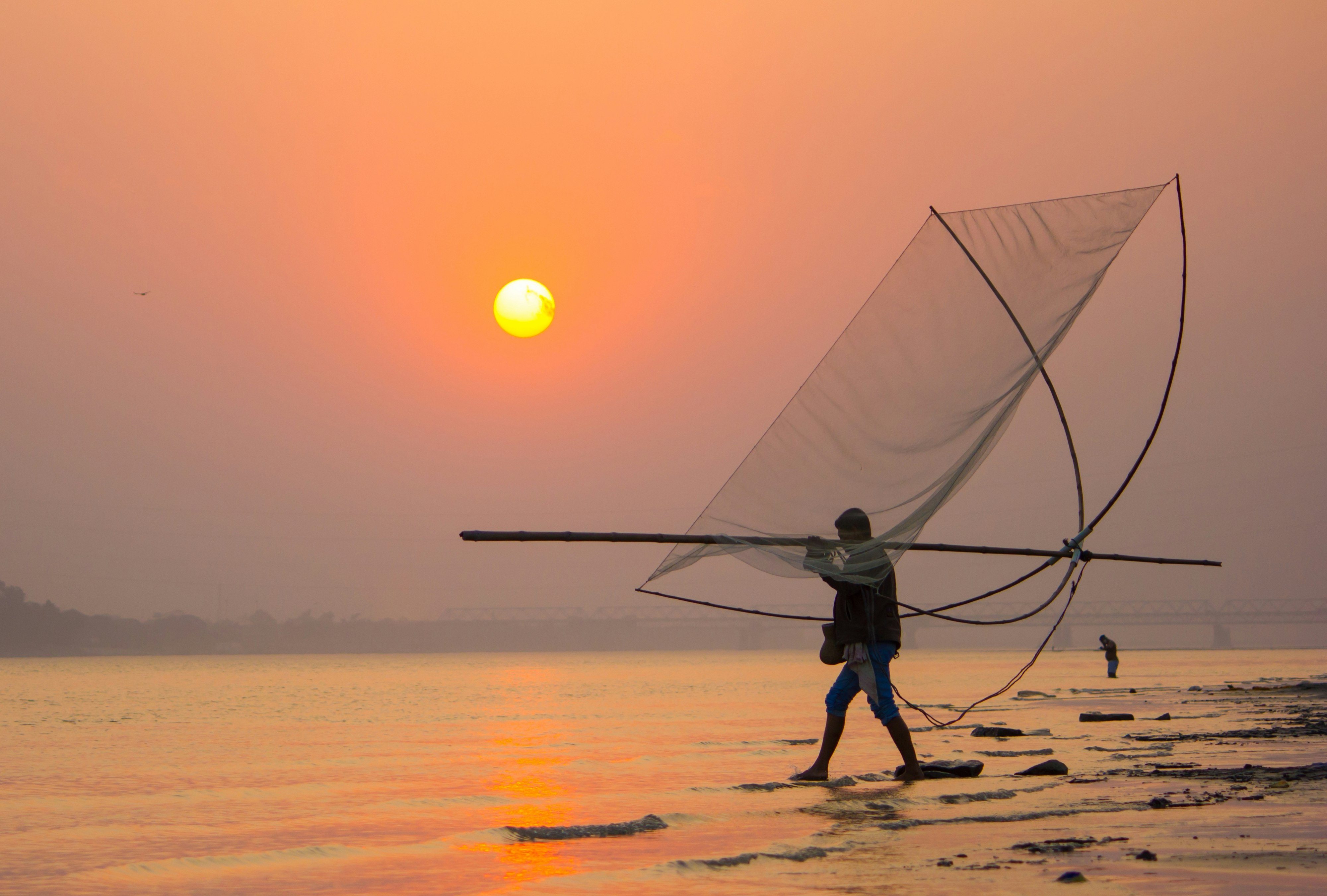 A fisherman, carrying a large net over his shoulder, wades into a river in Assam, with the low sun bathing the scene in an orange hue