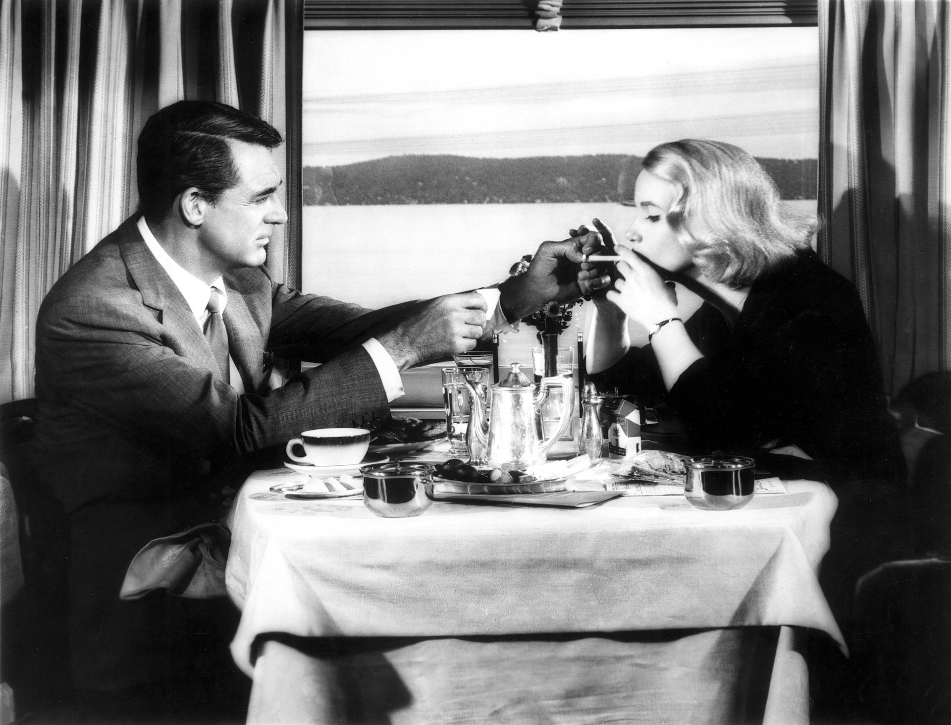 A black and white film still shows Cary Grant in a grey flannel suit lighting a cigarette for Eva Marie Saint in a black dress at the table of a train dining car dressed in white linen with a silver coffee service in Alfred Hitchcock's film 'North by Northwest' 