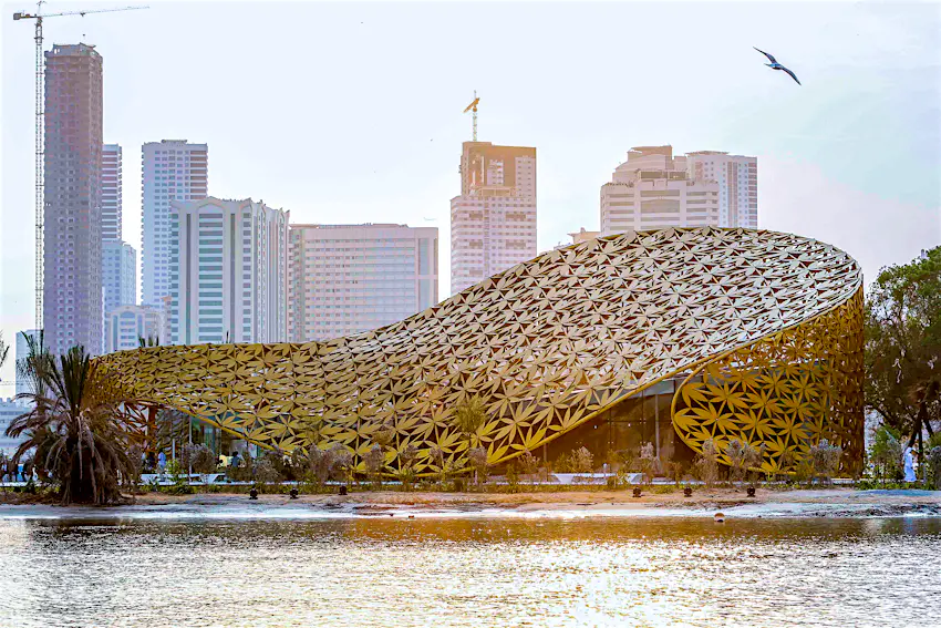 butterfly_house_sharjah.jpg A face-on view of a gold-coloured, undulating modern building. The curving facade is an open lattice; the fretwork is a pattern of palm leaves.