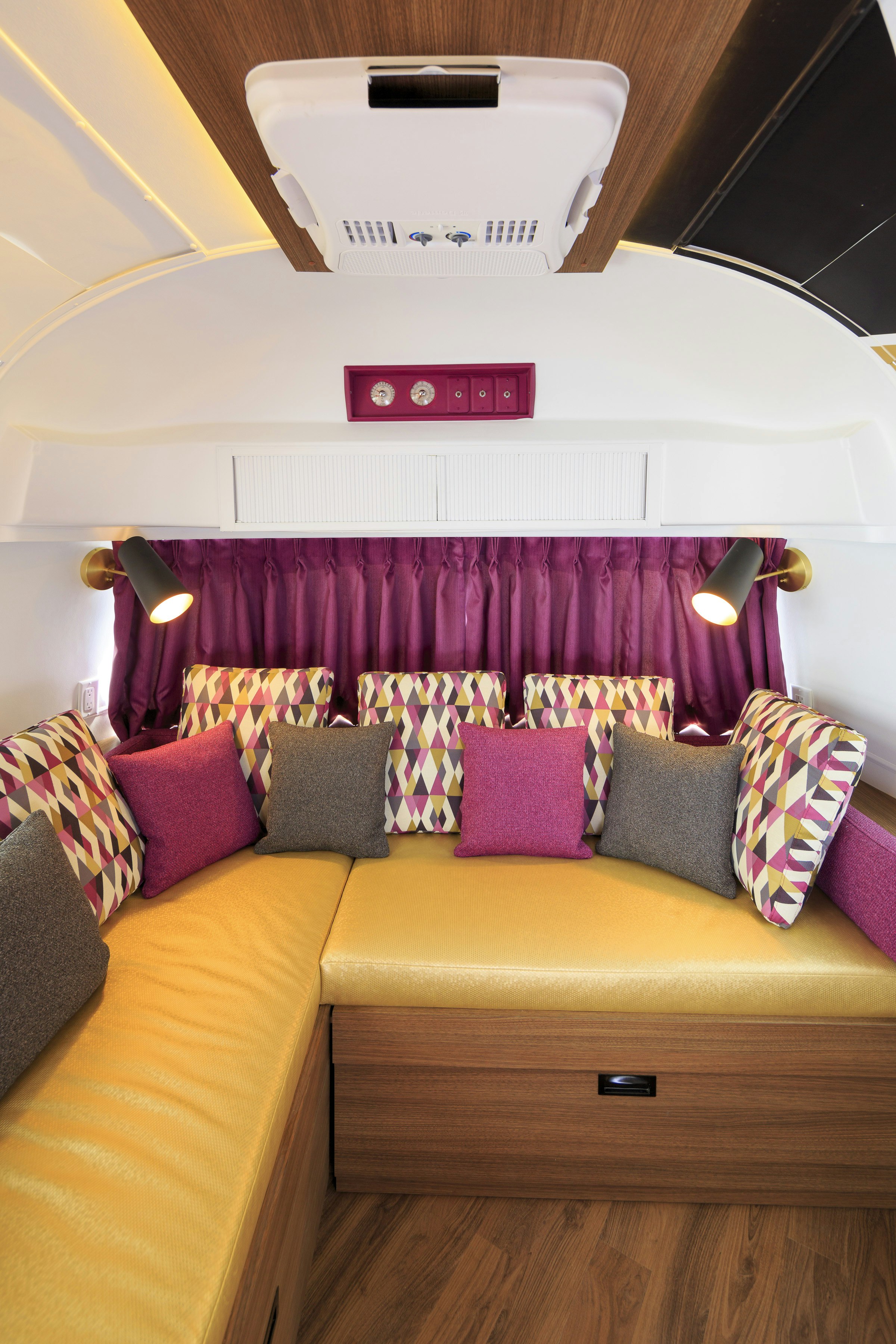 The colorful interior of the "Zedstream" Airstream trailer at Hotel Zed in British Columbia is decorated with warm wood, yellow vinyl seats, and magenta curtains that coordinate with pillows upholstered in pink, green, purple, and white harlequin patterns. 