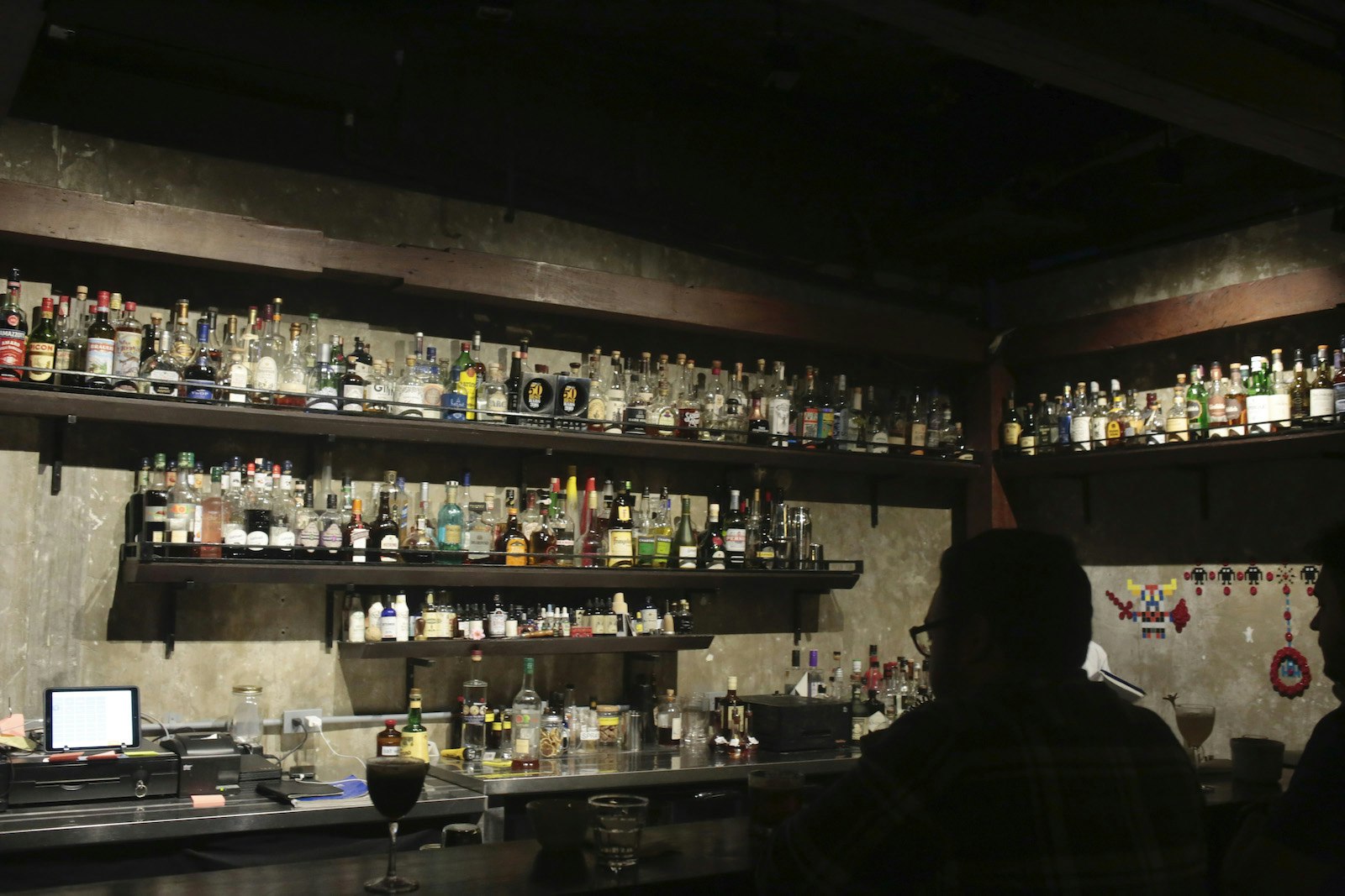 Dark room with a silhouette at the bar and three shelves of bottles behind the bar