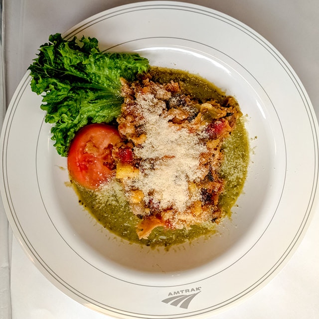 A white plate with the Amtrak logo on the edge in grey is filled with a Mexican fusion dish of elote sprinkled with finly shredded queso blanco, on a bed of verde sauce, with a bright red tomato and leaf of romaine lettuce as a garnish.
