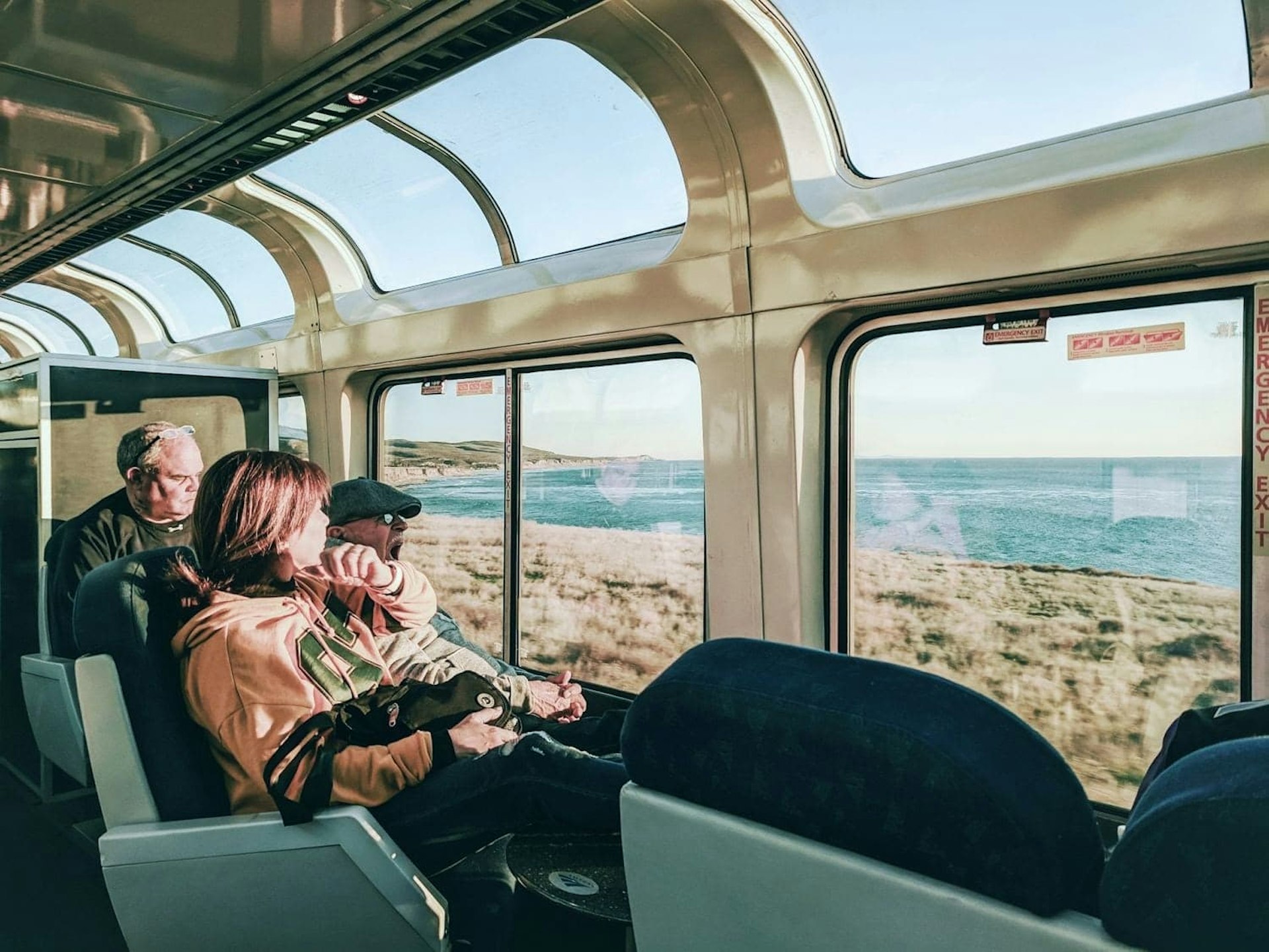 A trio of passengers sits in the viewing lounge car on the Coast Starlight route as it passes through Paso Robles. One older gentleman yawns as he admires the scenery. A woman with short dark hair is just turned away from the camera. A third man sits behind the yawning gent staring contemplatively out the window.