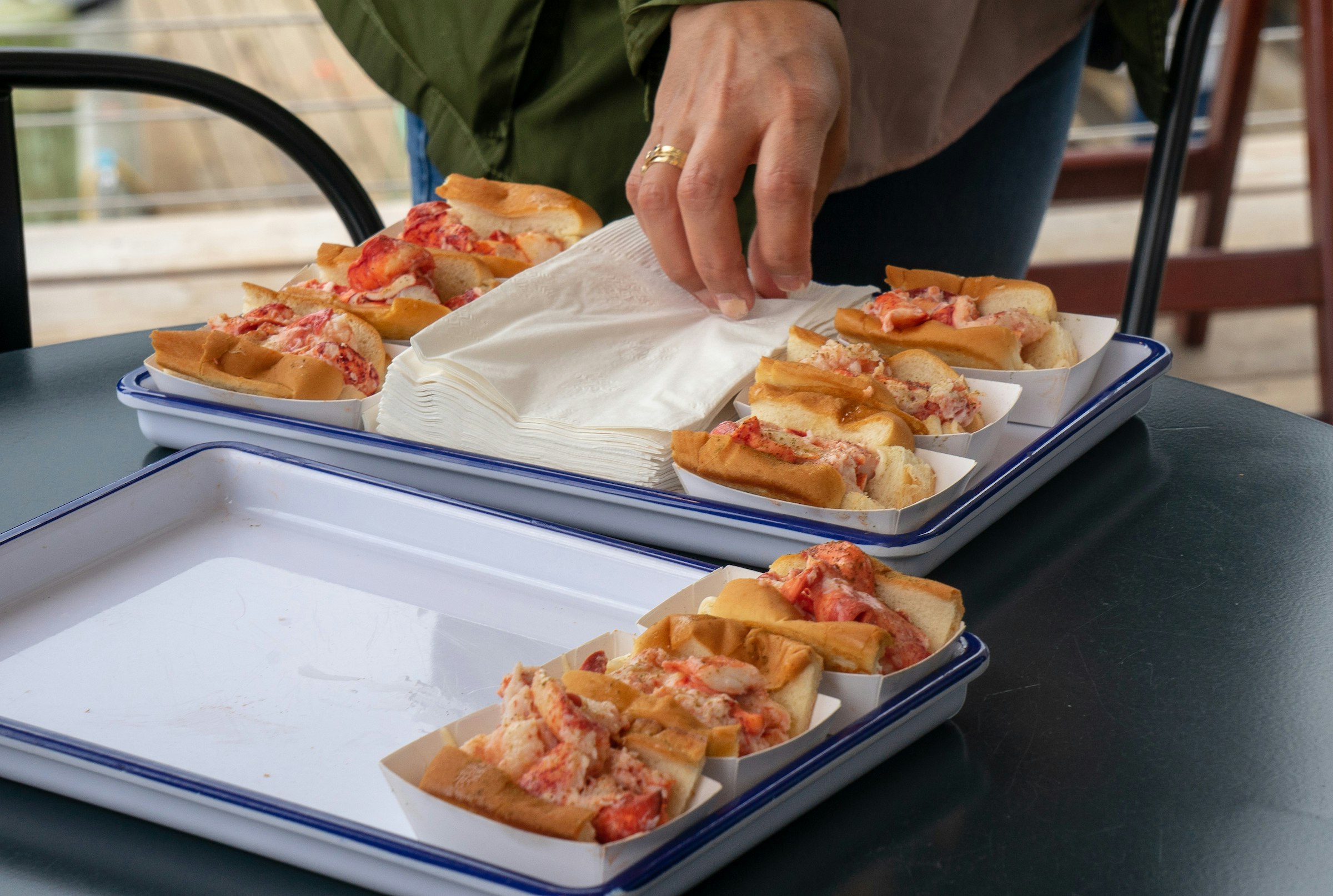 A hand wearing a gold wedding band reaches for a napkin from a pile set between two rows of Maine Lobster Rolls at Luke's in Portland. The rolls are in small paper trays and are heaped with bright red and pink lobster meat in white and blue enamel trays