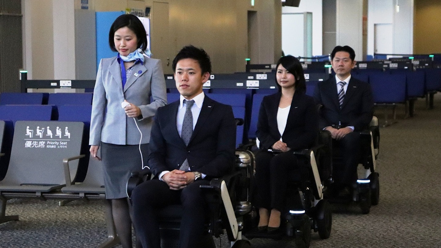 A line of three wheelchair users, accompanied by an ANA staffer, at Tokyo Narita International Airport
