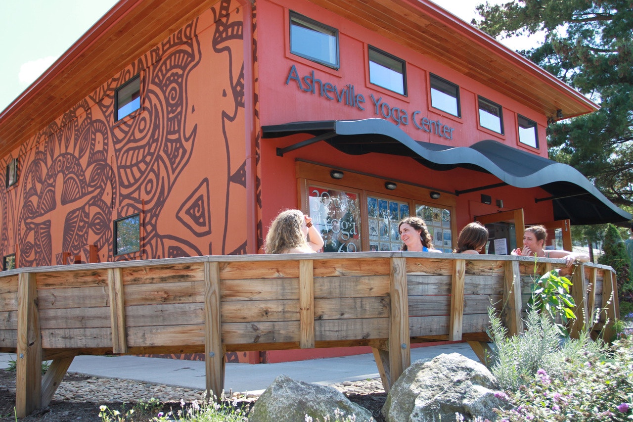 The exterior of the Asheville Yoga Center is a warm, deep red with a wavey black metal awning and five rectangular windows. A deep eave is lined with warm wood. On the left side of the building is a rown mural over an orange facade with a swiling, natural pattern and the outline of humans in tree and warrior yoga poses. On wooden benches in the foreground sit four women laughing and chatting after a yoga class.