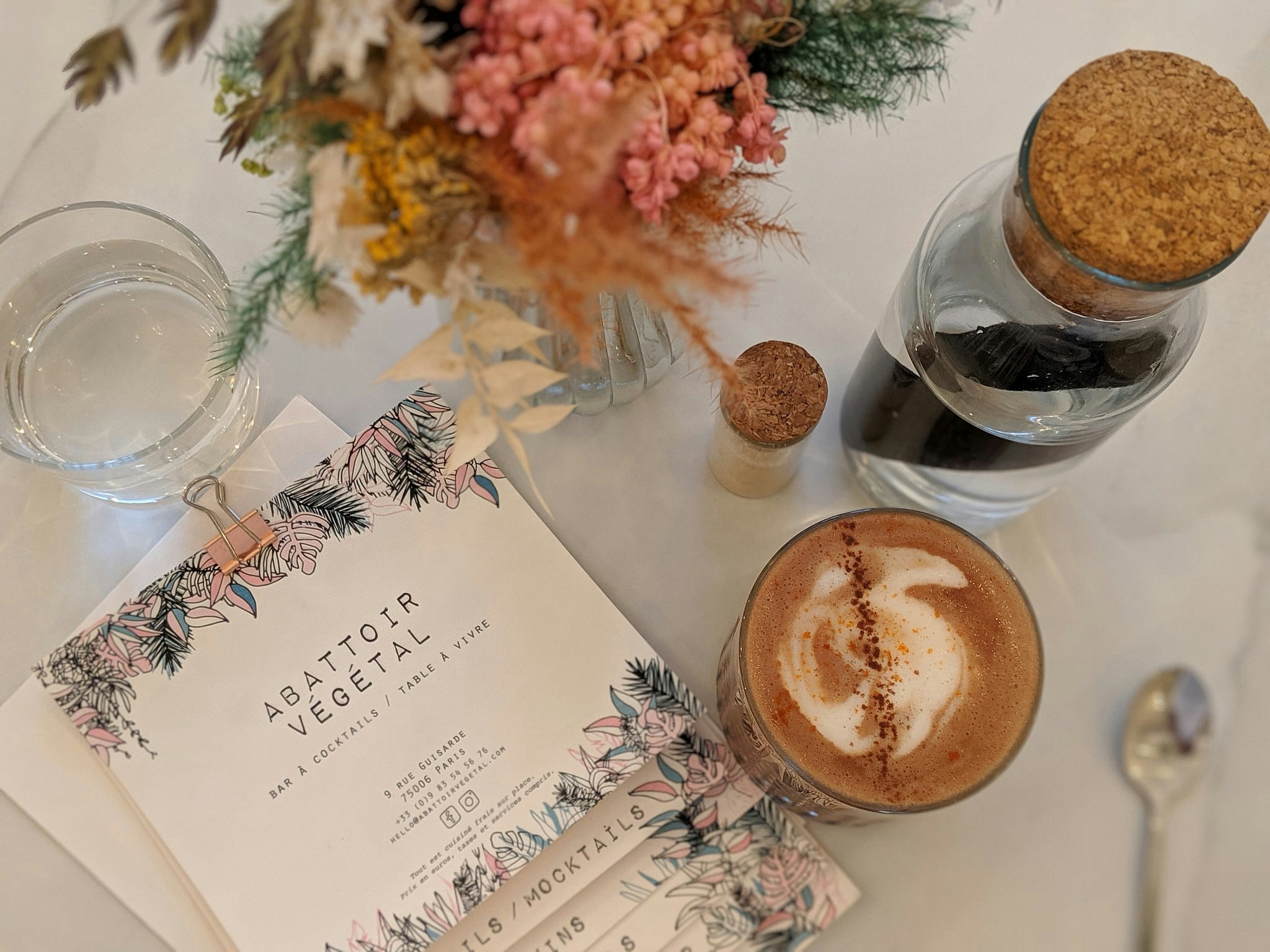 A latte on a white tablecloth next to corked water bottles, a floral display and a menu with a floral design.