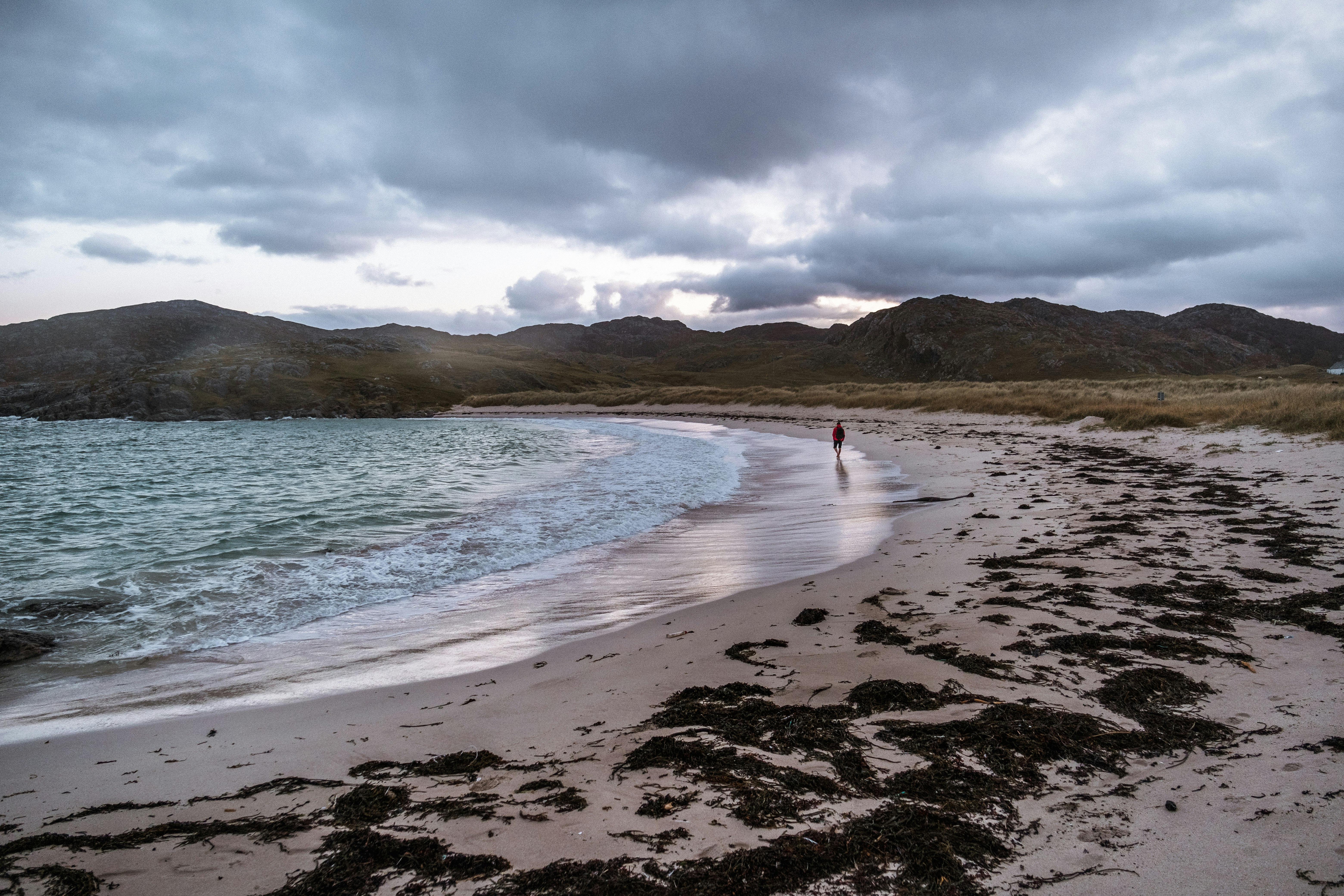 A man in a red jumper walks along an otherwise empty Achmelvich Beach.