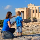 A woman kneeling next to a young boy points to the Acropolis in the distance in Athens.