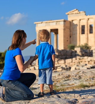 A woman kneeling next to a young boy points to the Acropolis in the distance in Athens.