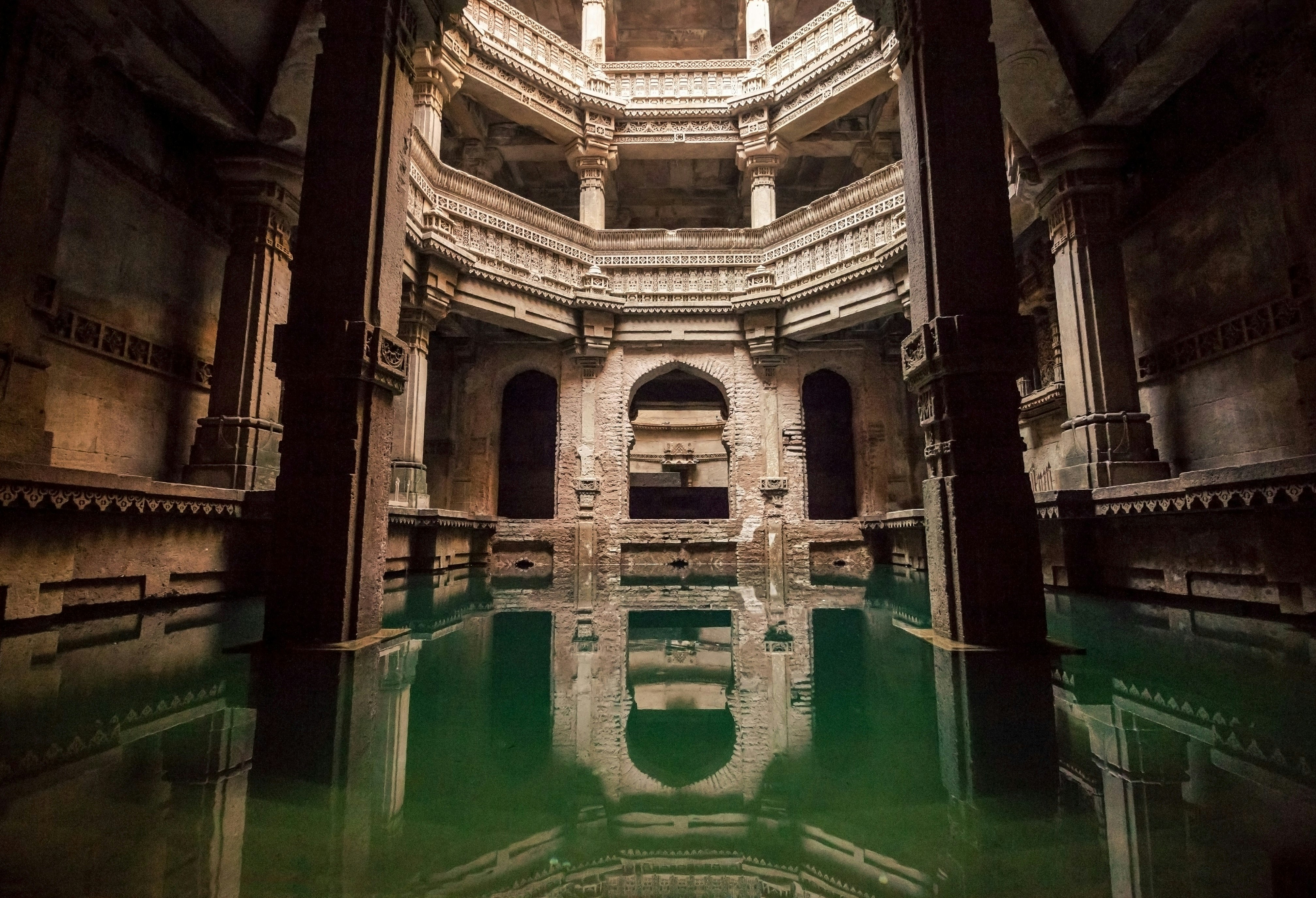 The interior of Adalaj Vav stepwell. The image is taken on the bottom of the stepwell, with the floor submerged in turquoise water. Light shines down from above, indicating it is the main shaft of the well.