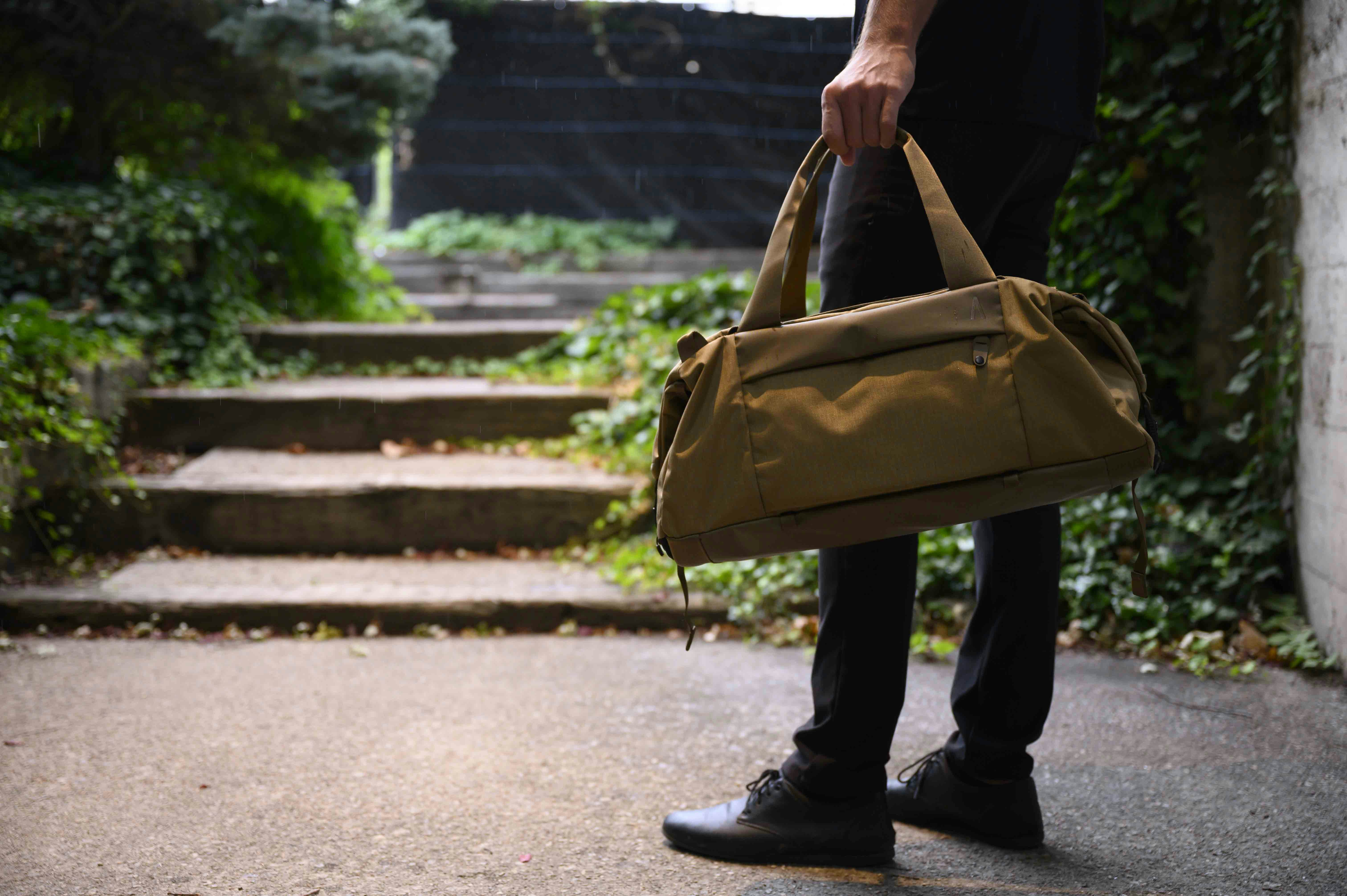 man in black holding Boundary Supply's Aegis duffel in brown, in front of steps with ivy on each side