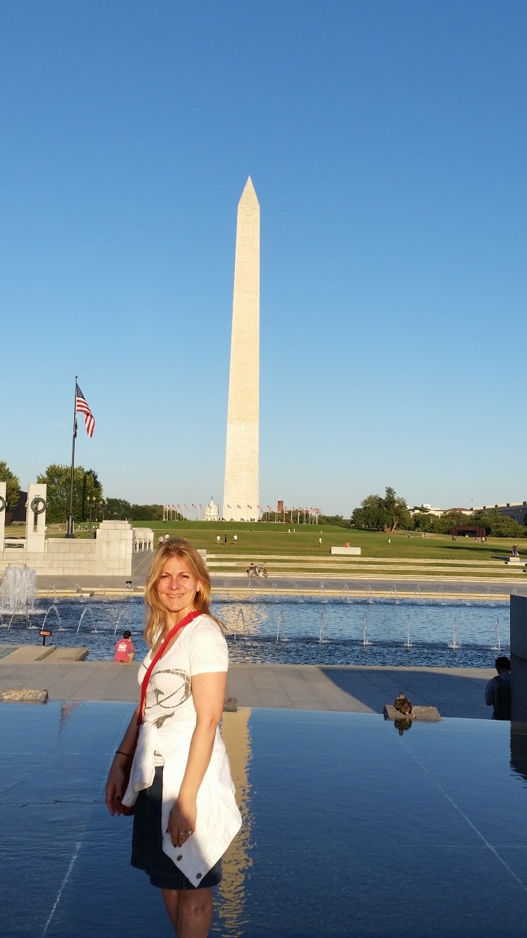 A woman is standing in front of the Washington Monument. She is standing side-on, and smiling at the camera. The monument is set against a bright blue sky.