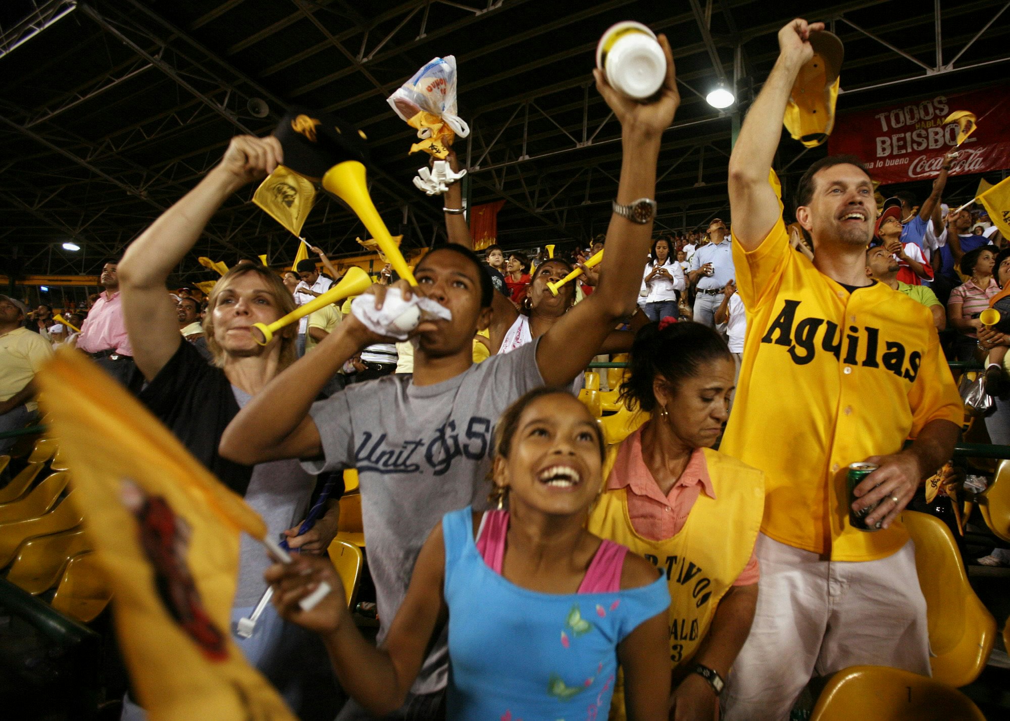 A group of people cheer (some with plastic horns) for the Aguilas baseball team in the Dominican Republic  