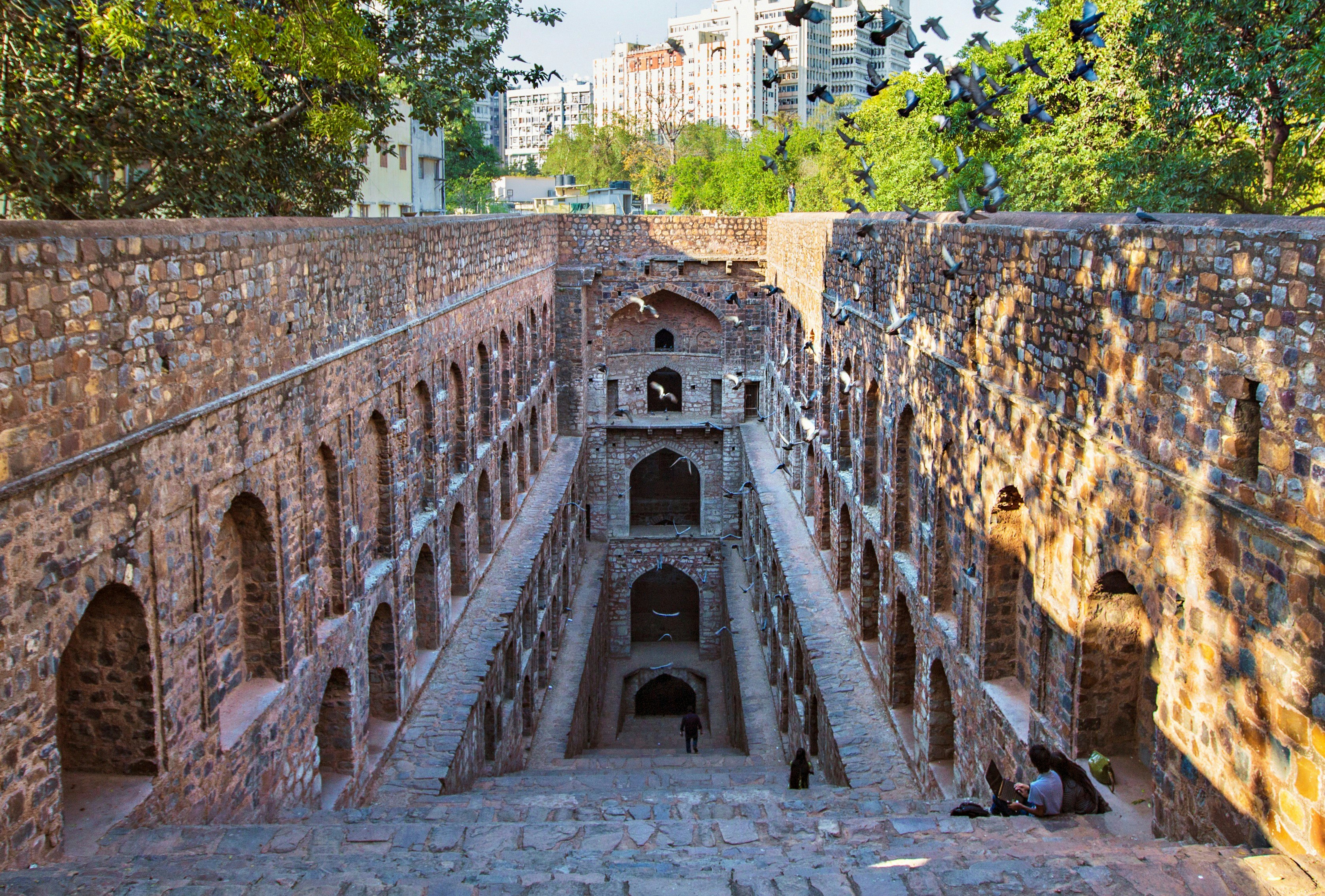 The stone Agrasen Ki Baoli stepwell in Delhi. A staircase descends downwards, enclosed by a vertical wall on all other sides, which are adorned with shallow arches. A few people sit on the steps, while, back on ground level, some trees and buildings are visible around the stepwell.