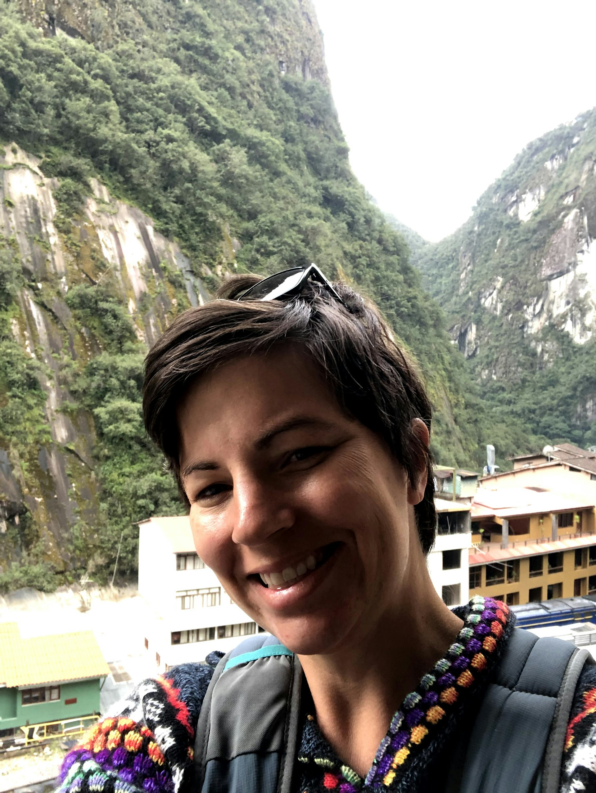 A woman wearing a colorful woven sweater smiles at the camera. In the background are tall green-covered mountains and small buildings.