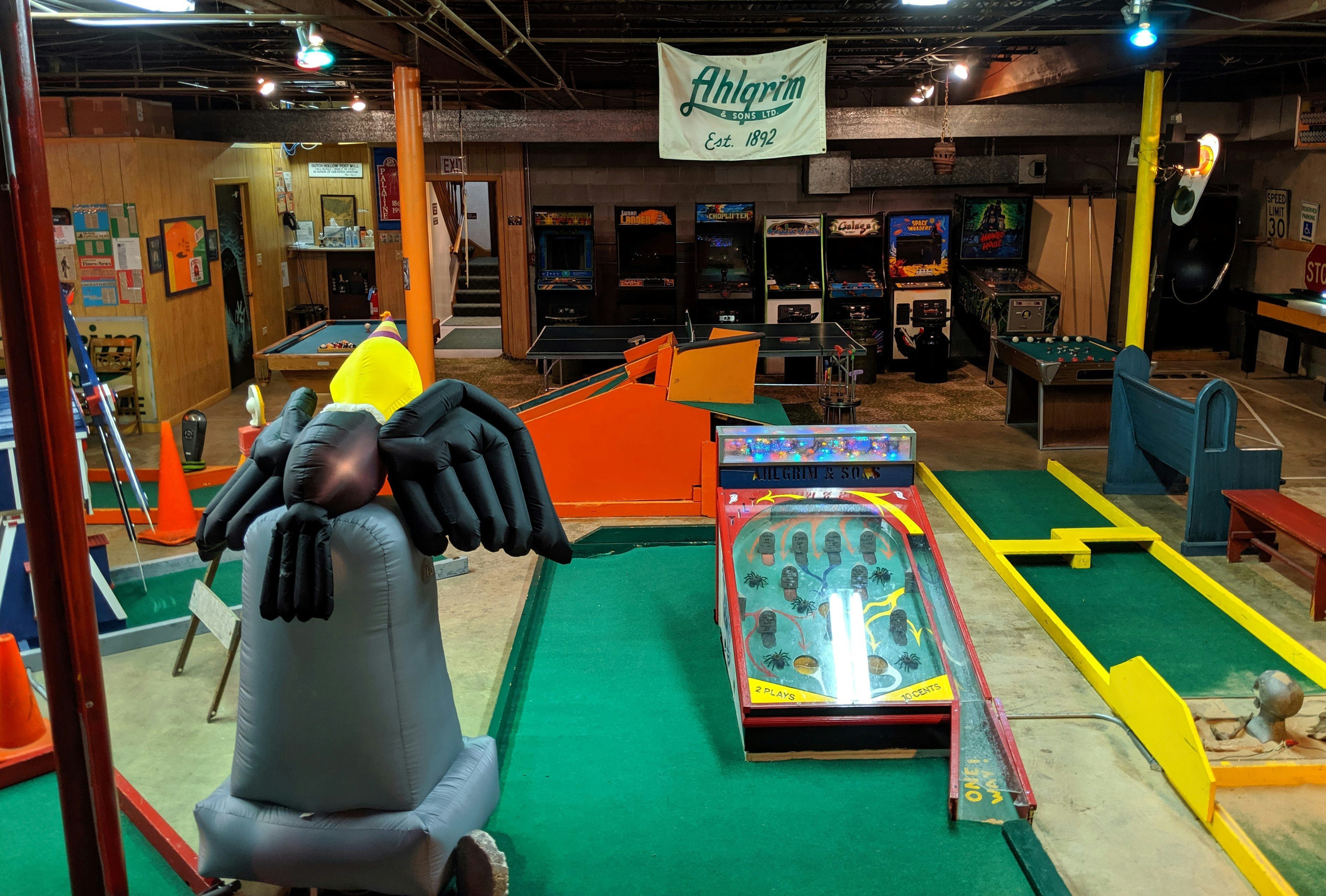 Ahlgrim Funeral Services mini golf course, which is located in the businesses' basement. There are a handful of makeshift 'greens' scattered around the room, featuring obstacles like a pinball machine and traffic cones. A pool table and arcade machines are also visible.