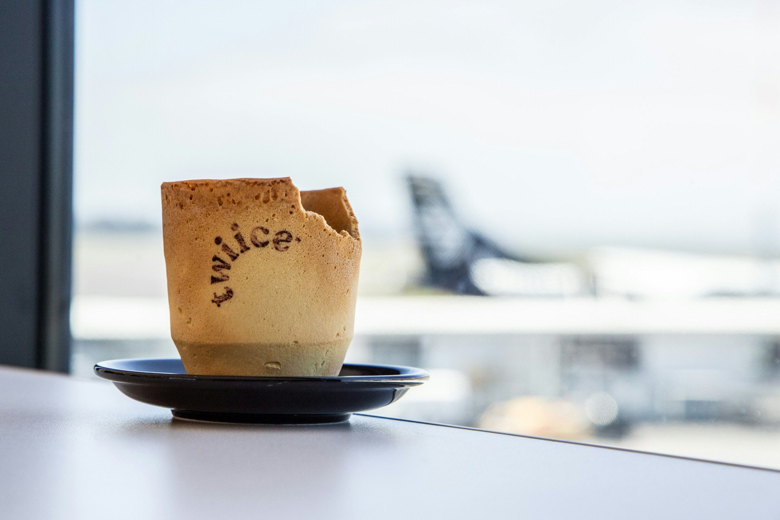 A biscuit cup with the Twiice logo sitting on a table in front of a window through which we can see and Air New Zealand aircraft
