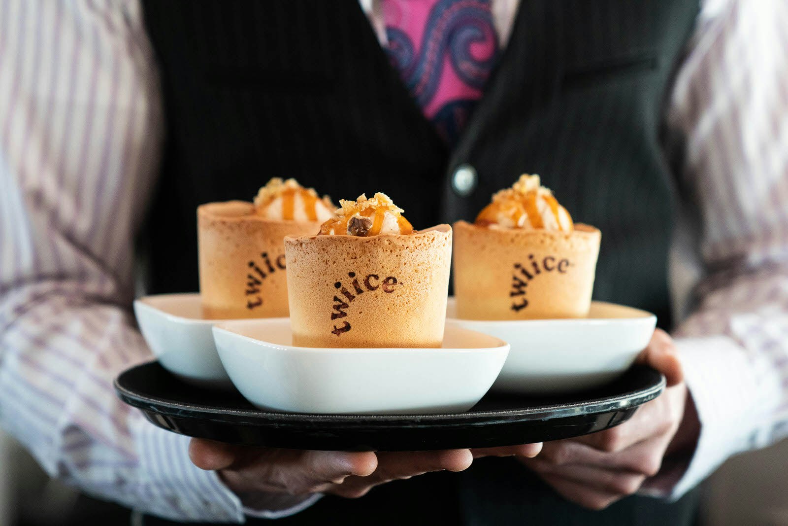 Three edible biscuit cups carried on a tray by someone wearing an Air New Zealand uniform. The biscuit cups are branded with the Twiice logo and have a whipped cream topping