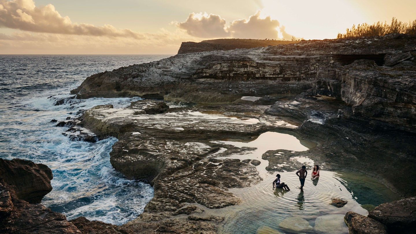 Three people wading/sitting in coastal tide pools as the sun sets behind a cliff