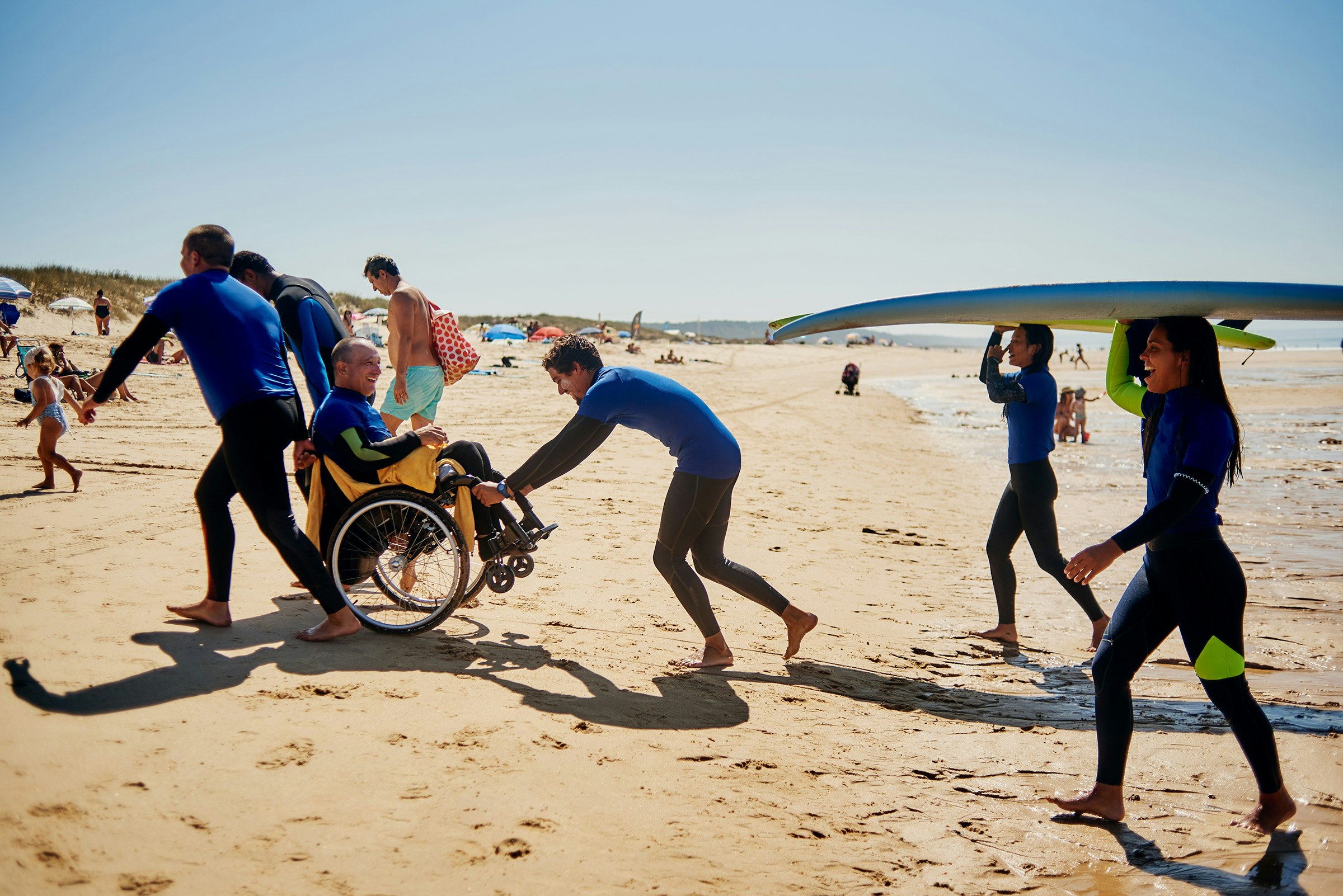 A group of people smiling on a beach with surfboards, one man is in a wheelchair